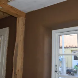 Ściana po tynkowaniu gliną w odcieniach brązu, widoczny drewniany element konstrukcyjny i okno z widokiem na zewnątrz, gdzie osoba pracuje z taczką.