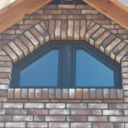 Trójkątne okno w elewacji z cegły klinkierowej, z drewnianą konstrukcją dachu w tle. Ciekawy układ cegieł wokół okna w kształcie łuku.