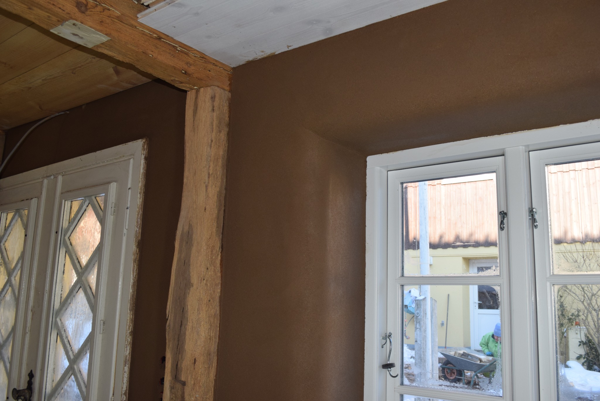 Ściana po tynkowaniu gliną w odcieniach brązu, widoczny drewniany element konstrukcyjny i okno z widokiem na zewnątrz, gdzie osoba pracuje z taczką.