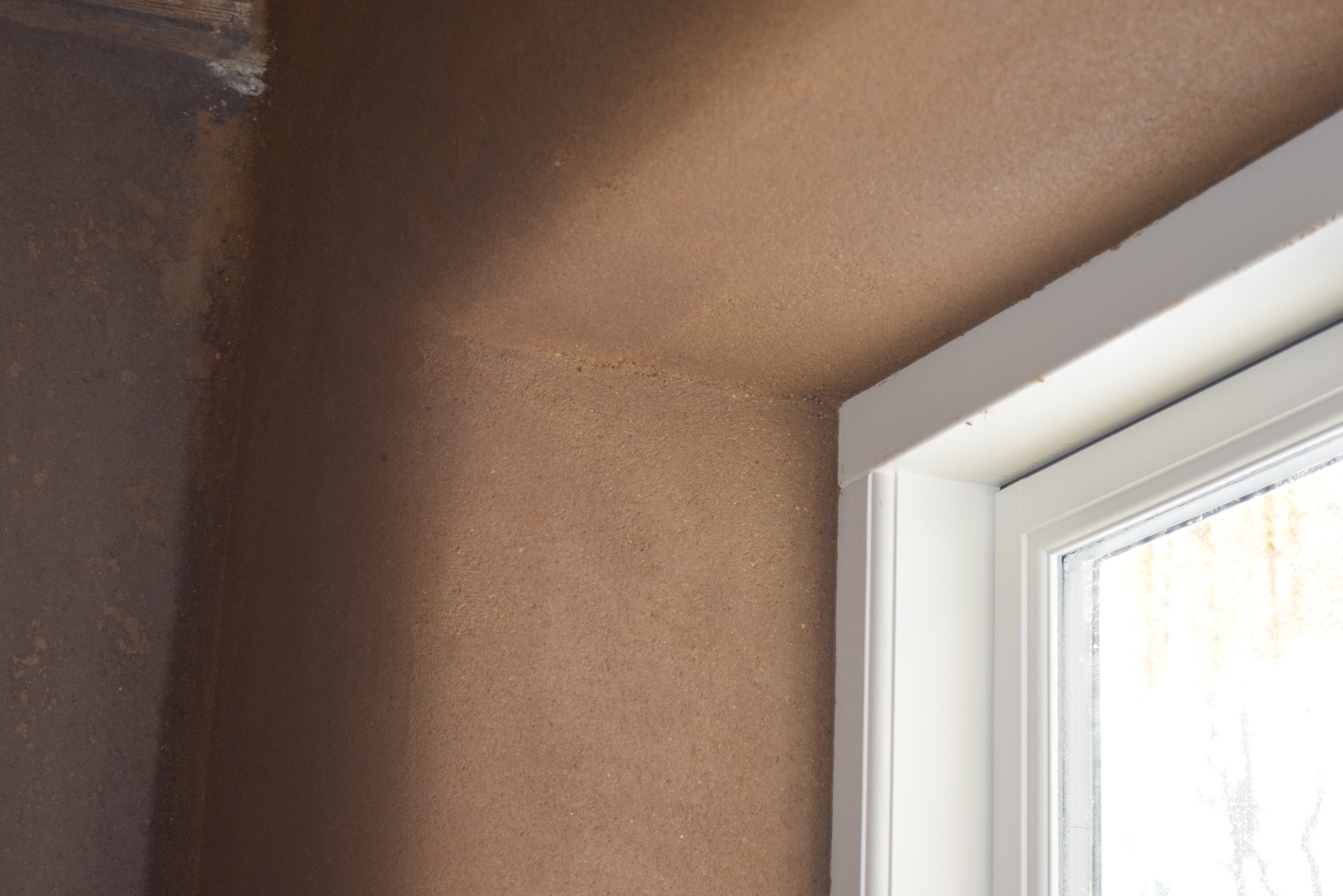 Surowa, brązowa ściana z fakturą ziarnistą, styka się z białą ramą okienną. Widoczny róg pomieszczenia z niejednolitą powierzchnią.