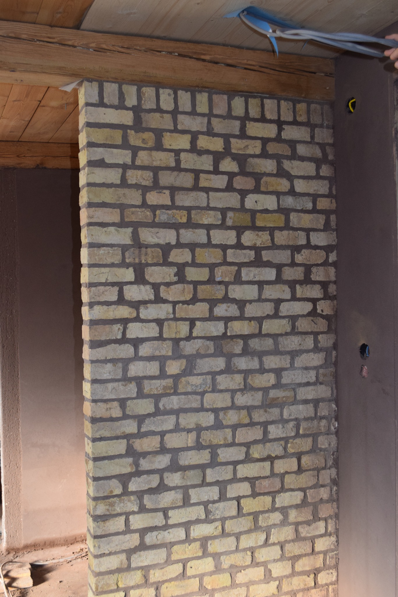 Murowana ściana z żółtej cegły w trakcie prac wykończeniowych. Widoczne przewody elektryczne i fragment ściany przygotowanej do tynkowania.