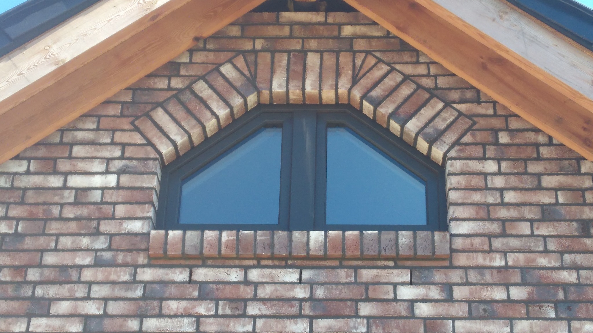 Trójkątne okno w elewacji z cegły klinkierowej, z drewnianą konstrukcją dachu w tle. Ciekawy układ cegieł wokół okna w kształcie łuku.