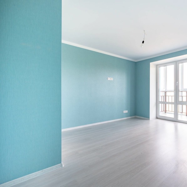 Pomalowane na turkusowo wnętrze pokoju z jasną podłogą i białymi listwami przypodłogowymi. Widoczne gniazdka elektryczne i balkon z białymi drzwiami.