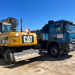 Żółta koparka CAT M313D i niebieska ciężarówka z logo firmy z Lidzbarka Warmińskiego na placu budowy pod błękitnym niebem.