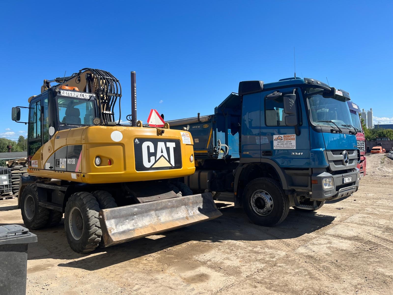 Żółta koparka CAT M313D i niebieska ciężarówka z logo firmy z Lidzbarka Warmińskiego na placu budowy pod błękitnym niebem.