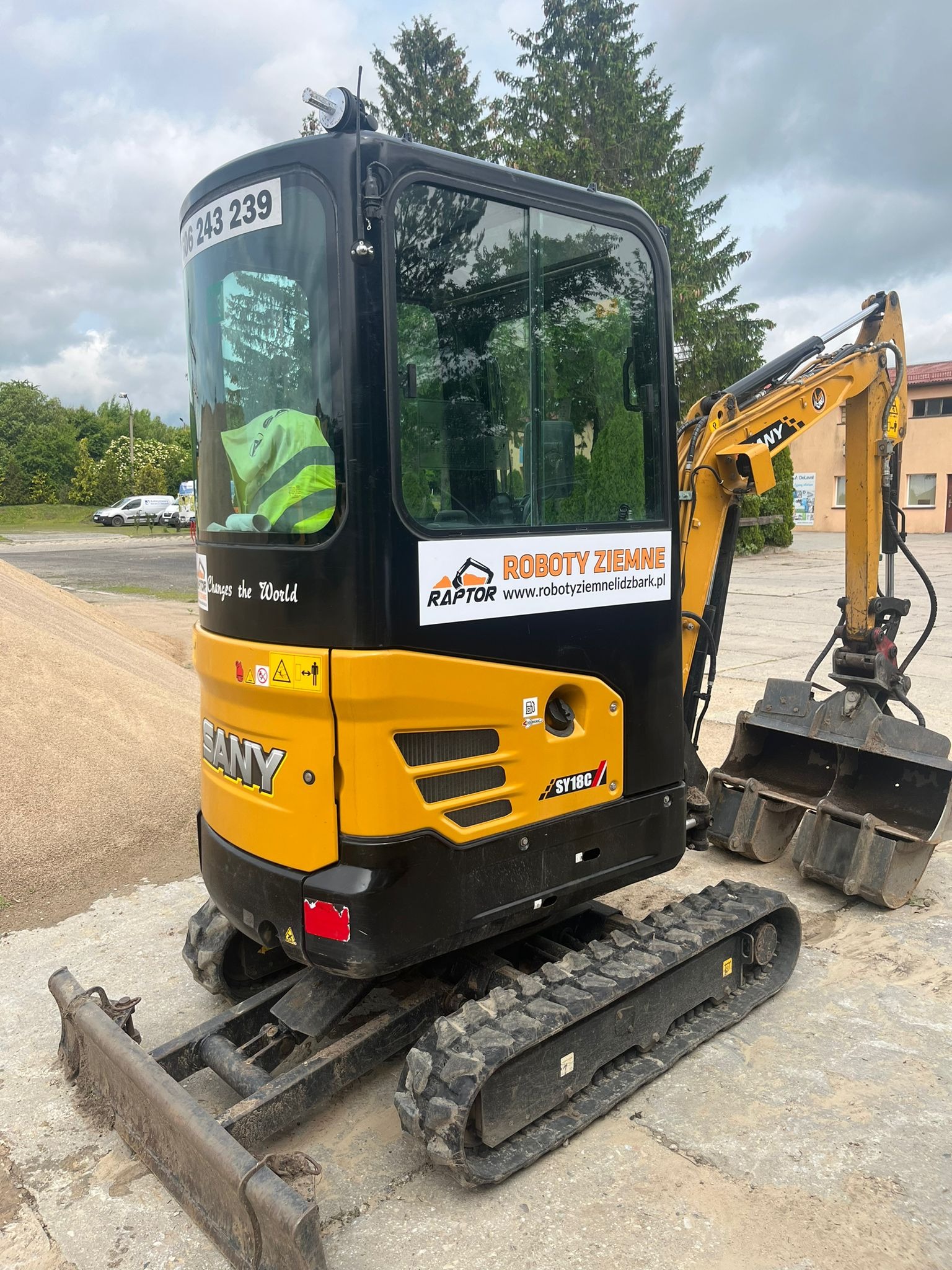 Żółto-czarna mini koparka SANY SY18C z łyżką, z naklejką 'Roboty Ziemne' i numerem telefonu, stoi na betonowej powierzchni. W tle widać stertę piasku i zieleń.