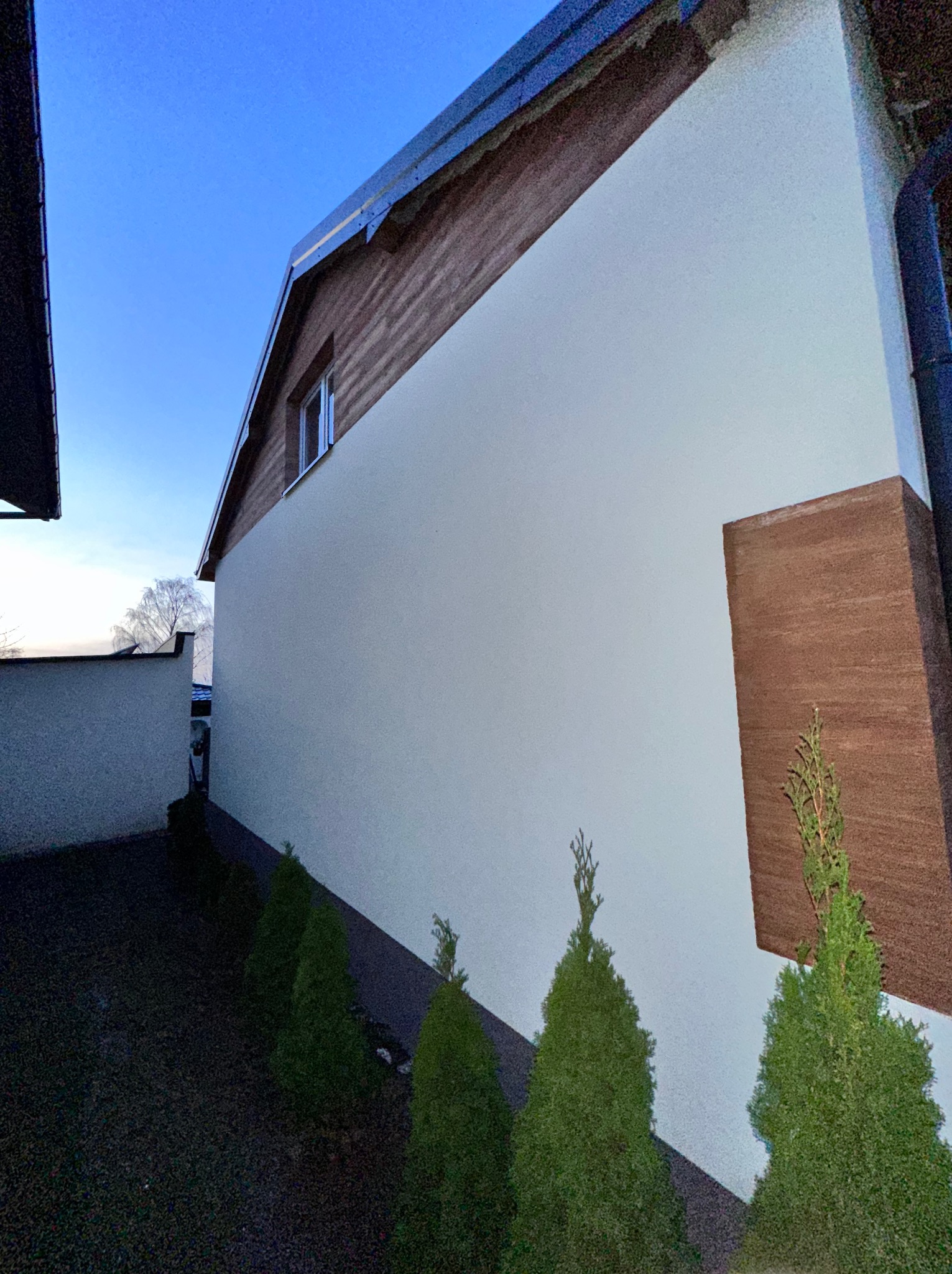 Elewacja domu jednorodzinnego z drewnianymi akcentami w Głownie. Jasna ściana kontrastuje z ciemnym dachem i elementami dekoracyjnymi. Widoczne tuje.