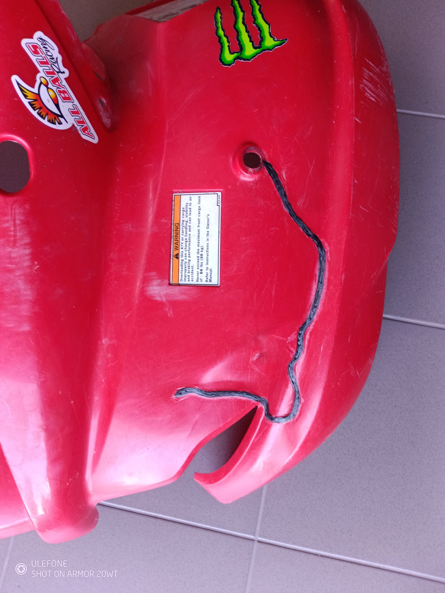 Czerwony plastikowy element z naklejkami (All Balls Racing, Monster) i czarnym szwem. Widoczne ślady użytkowania i ostrzeżenie o wadze ładunku.