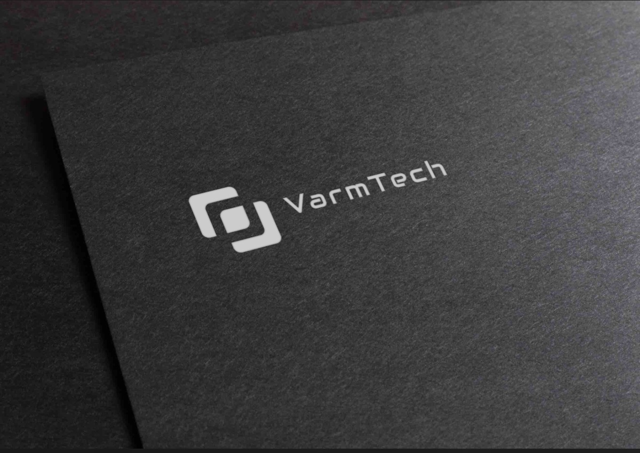 Minimalistyczny, biały logotyp 'VarmTech' z geometrycznym symbolem na teksturowanym, ciemnoszarym tle, ujęty pod kątem, tworzący elegancki i nowoczesny wizerunek.