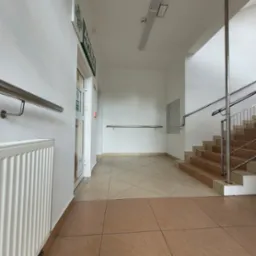 Jasne wnętrze budynku po malowaniu: białe ściany, grzejnik, schody z metalową barierką, płytki na podłodze. Widoczny fragment apteki.