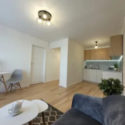 Jasne wnętrze mieszkania po odświeżeniu: białe ściany, drewniane panele, nowoczesna kuchnia i salon z szarą sofą. Widok og&oacute;lny, ciepłe oświetlenie.