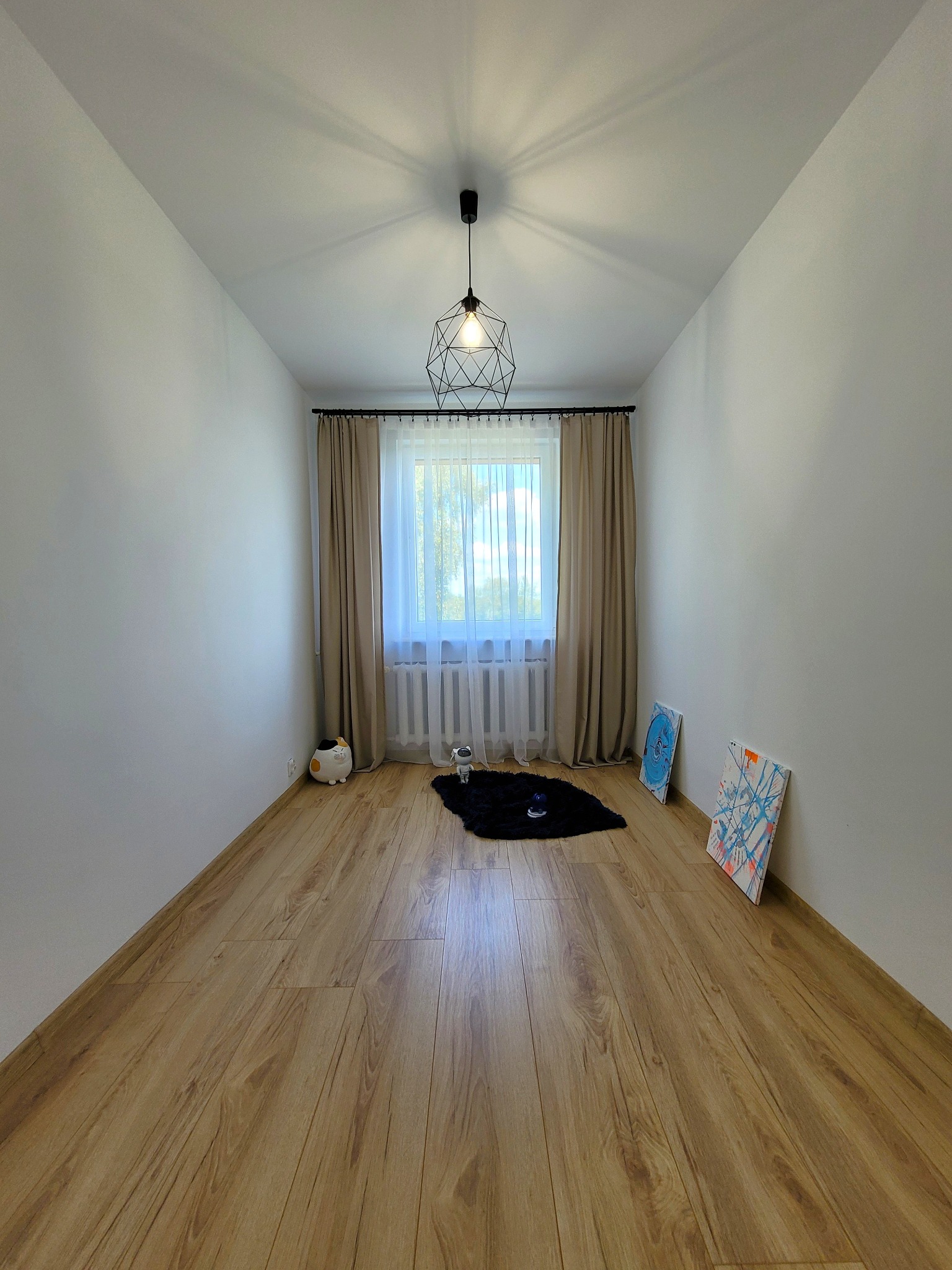 Pusty pokój po odświeżeniu: białe ściany, drewniana podłoga, okno z beżowymi zasłonami i lampa sufitowa. Dwa obrazy opierają się o ścianę.