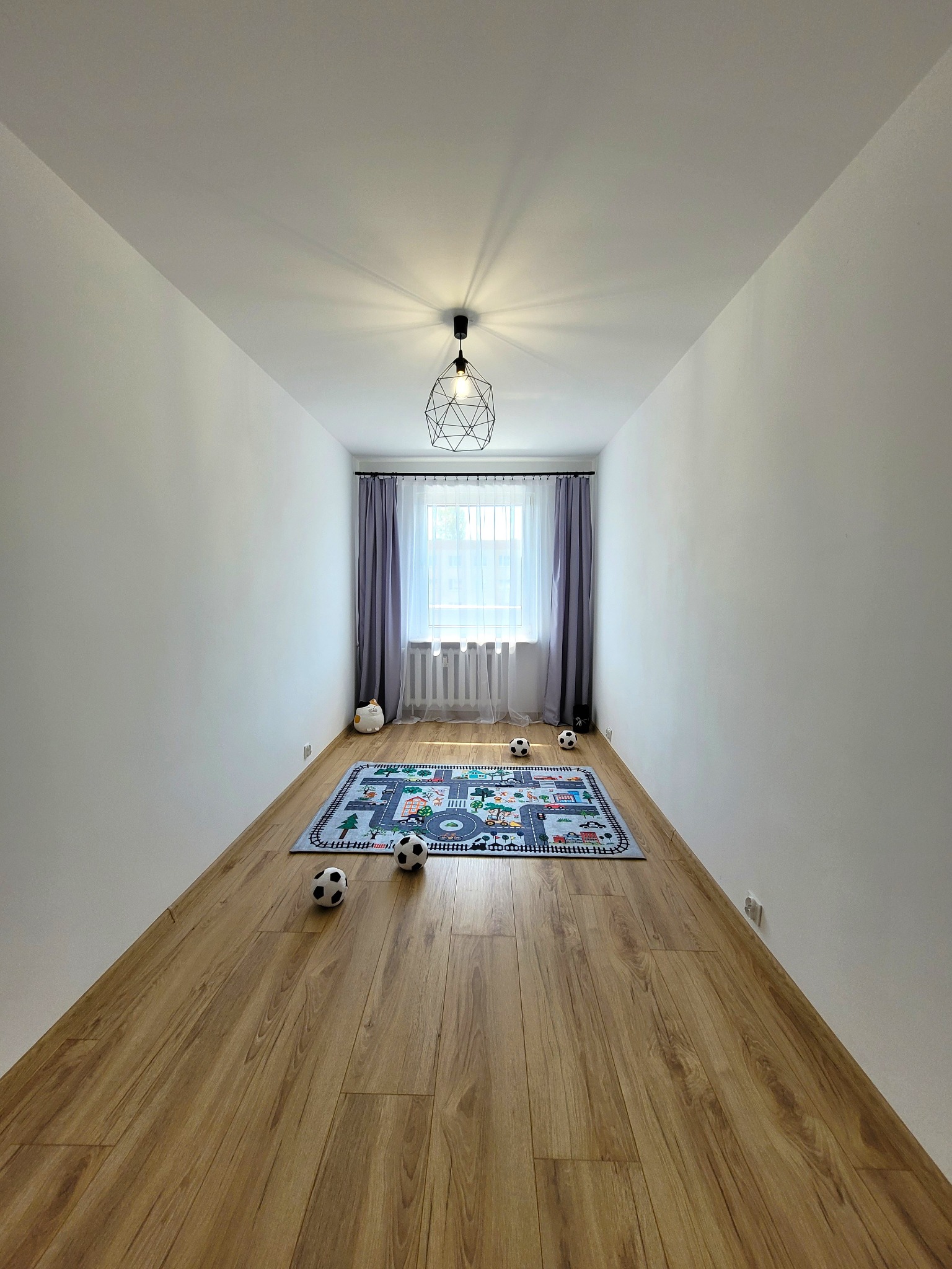 Pomalowane na biało wnętrze pokoju z drewnianą podłogą, dywanem z motywem ulic i oknem z zasłonami. Nowoczesna lampa sufitowa.