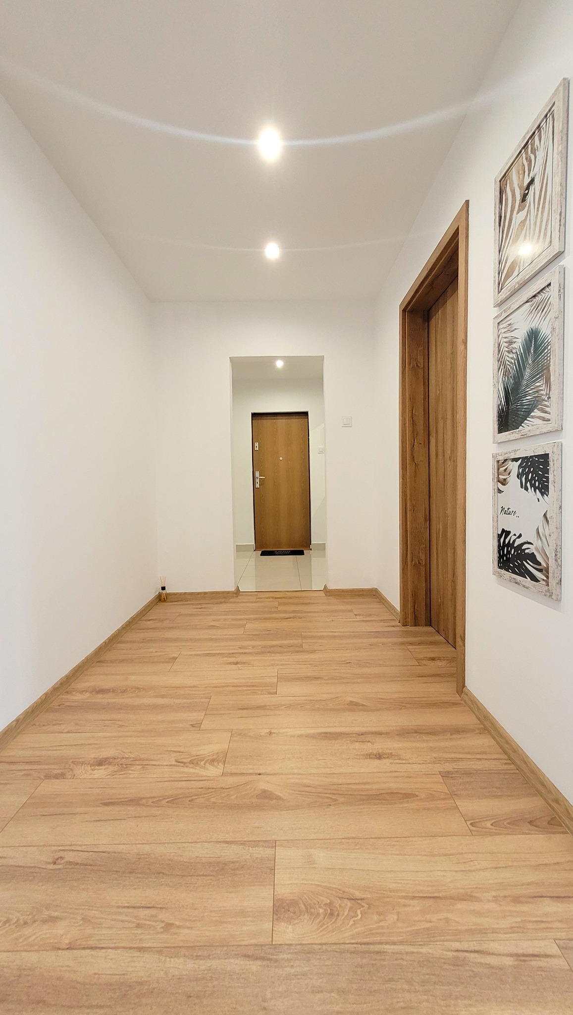 Jasny korytarz po malowaniu: białe ściany, drewnopodobna podłoga, drzwi z drewnianą ościeżnicą i dekoracje ścienne z motywami roślinnymi.