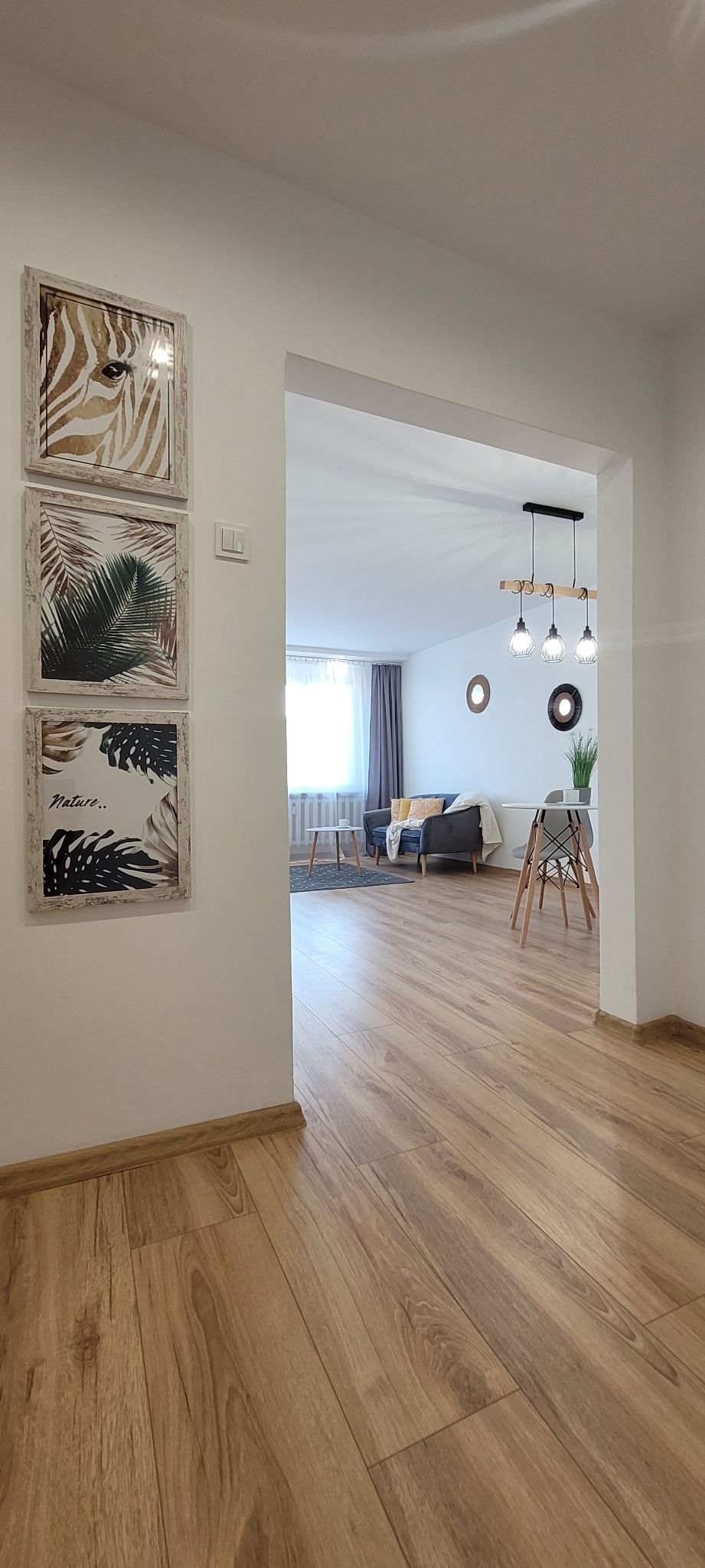 Wykończone wnętrze mieszkania z drewnianą podłogą, białymi ścianami i dekoracjami ściennymi. Widok na salon z szarą sofą i oknem.