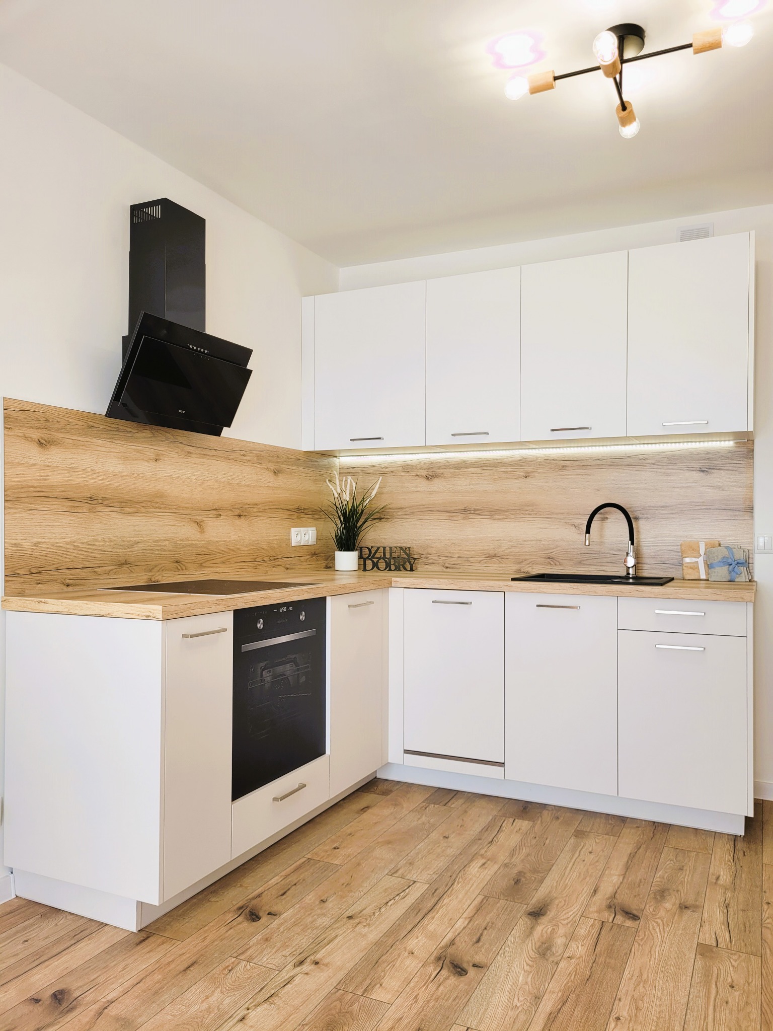 Jasna kuchnia z białymi szafkami i drewnianymi akcentami: blat, panel nad blatem, podłoga. Nowoczesny design z czarnym okapem i minimalistyczną lampą.