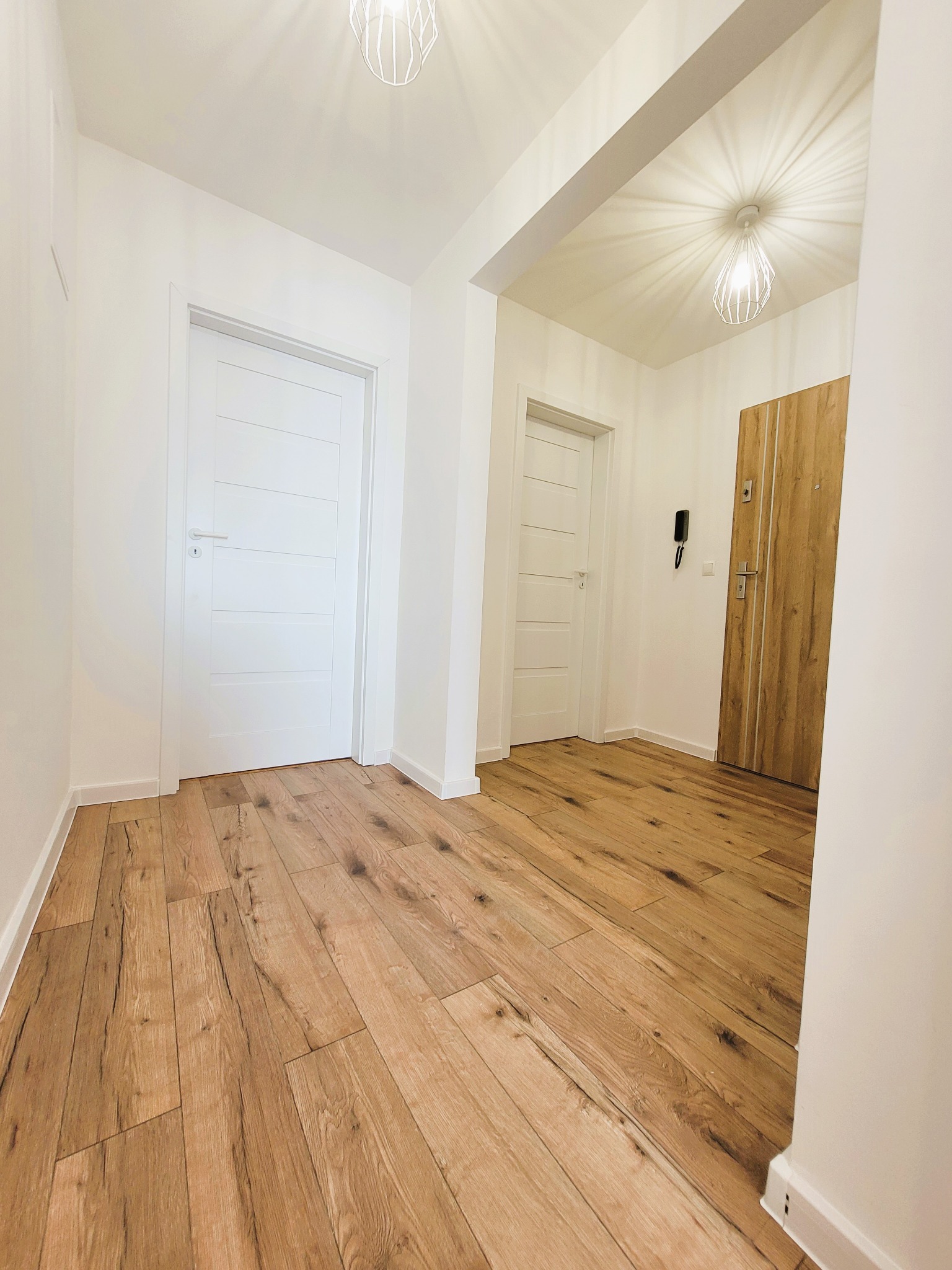 Jasne wnętrze mieszkania po remoncie: białe ściany, drewniana podłoga, minimalistyczne drzwi i nowoczesne oświetlenie. Widok na korytarz.