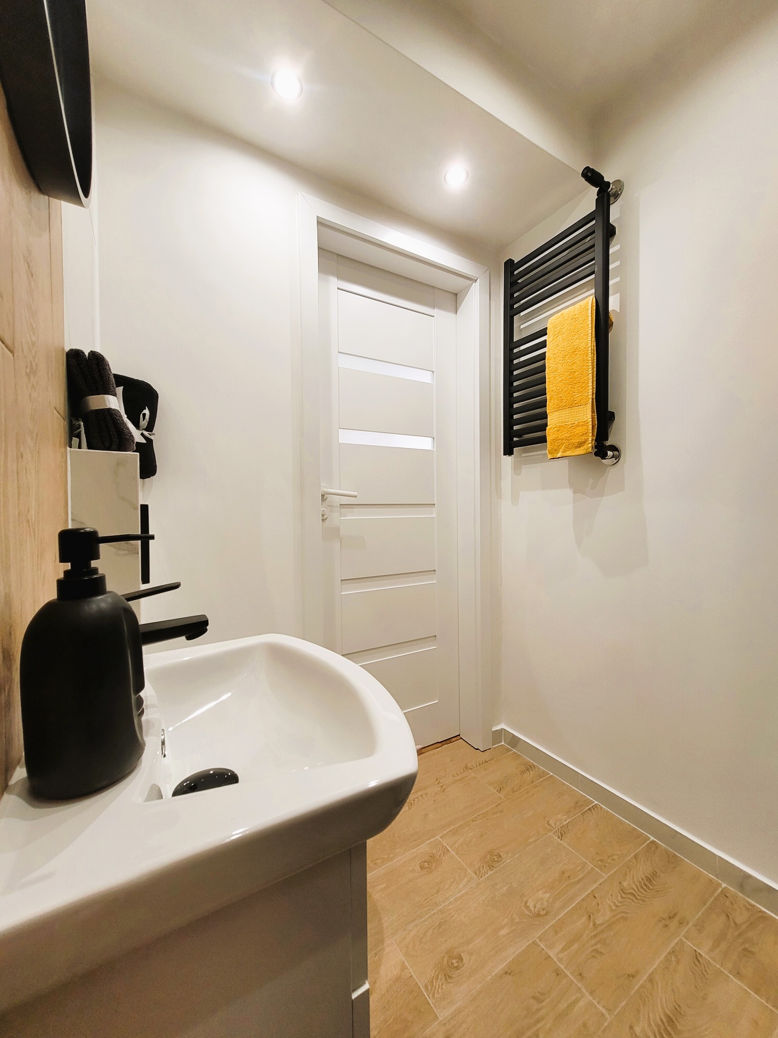Biała umywalka w nowoczesnej łazience z drewnopodobną podłogą i czarnym grzejnikiem z żółtym ręcznikiem. Ściany pomalowane na biało, minimalistyczny design.