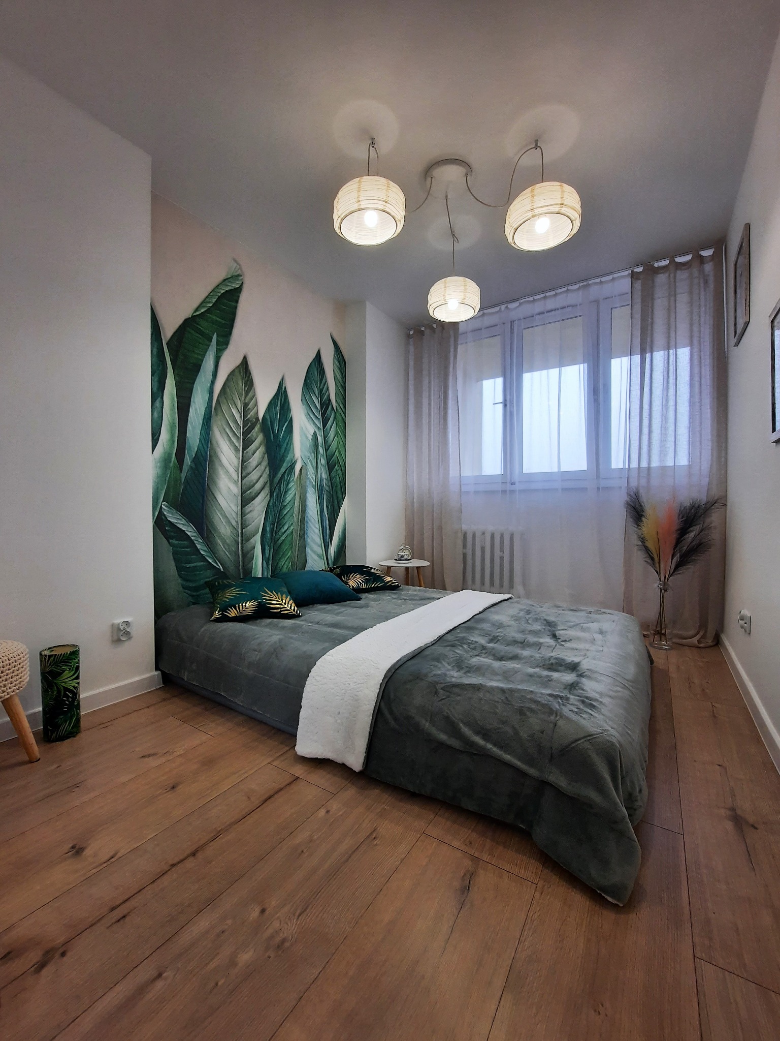 Sypialnia z fototapetą w tropikalne liście, drewniana podłoga, szare łóżko i lampy w stylu boho. Dekoracja wnętrza z akcentem natury i ciepłym oświetleniem.
