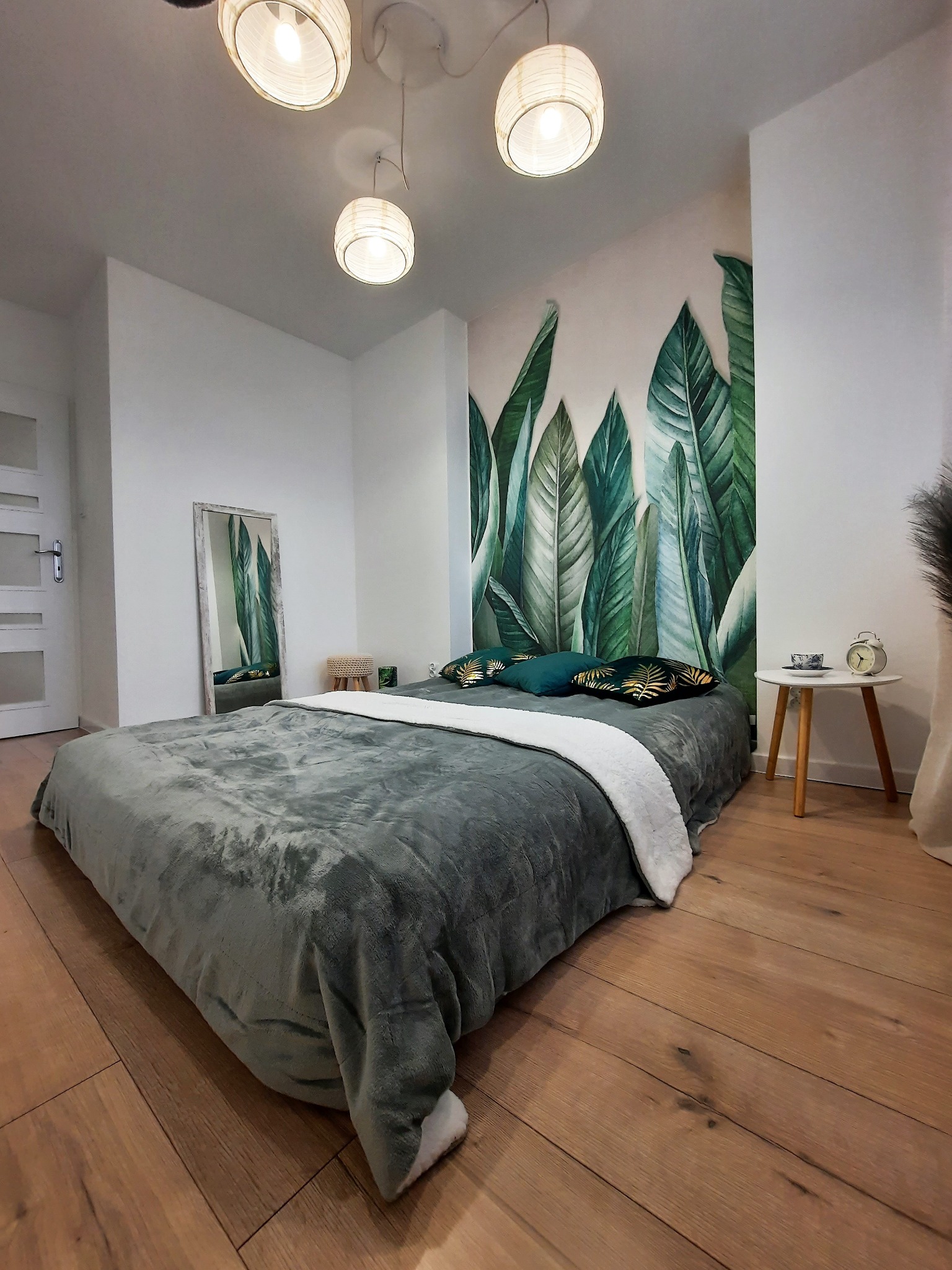 Pokój z łóżkiem, ścianą z motywem liści i drewnianą podłogą. Trzy lampy wiszące oświetlają wnętrze. W tle lustro i stolik nocny z zegarem.