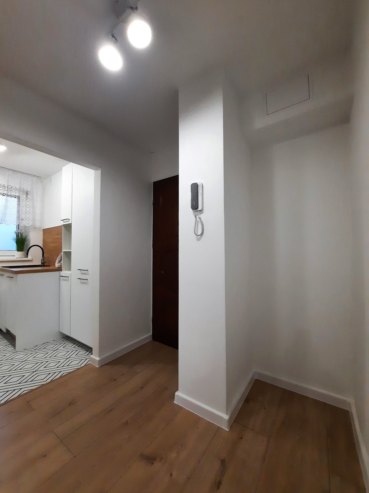 Białe ściany po odświeżeniu w mieszkaniu, widoczny fragment kuchni z białymi szafkami i drewnianym blatem, brązowe panele na podłodze i domofon na ścianie.