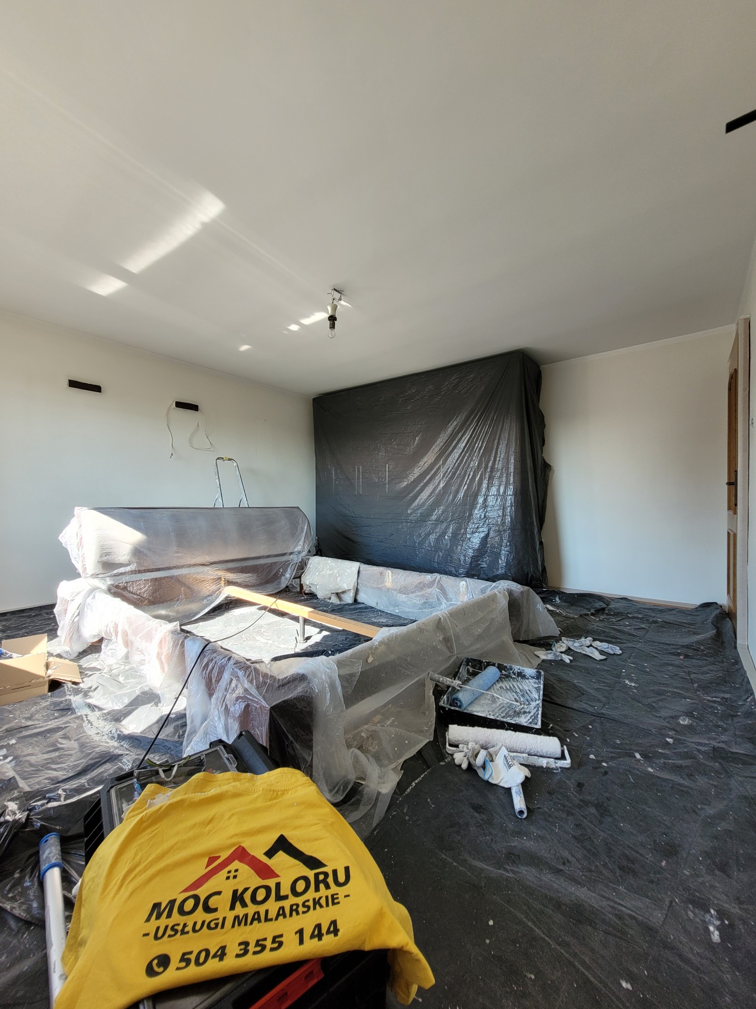 Przygotowania do malowania mieszkania w Katowicach. Meble zabezpieczone folią, podłoga przykryta, widoczny wałek i kuweta malarska oraz koszulka firmowa.