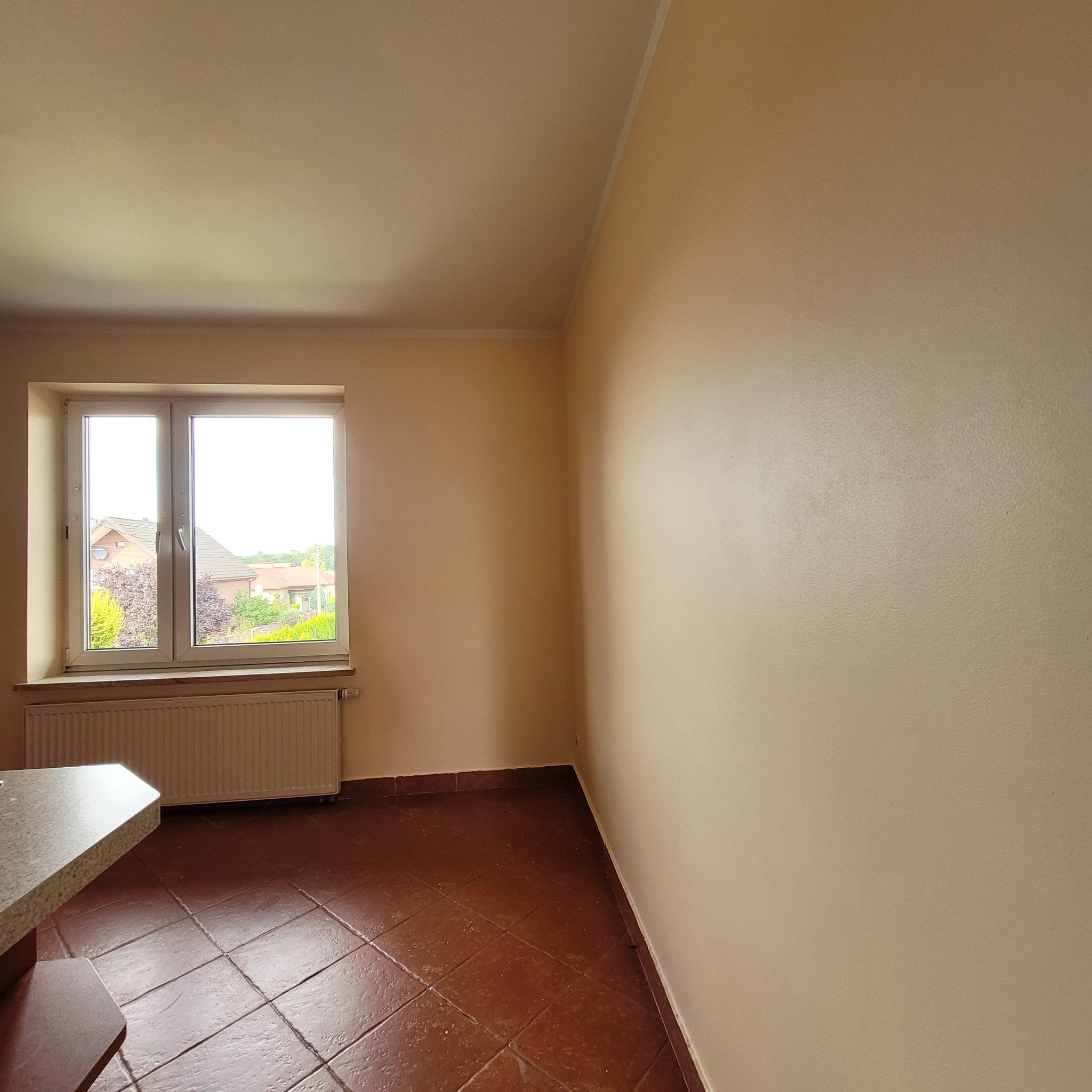 Pomalowane na beżowo wnętrze pokoju z oknem i brązową podłogą z płytek. Widoczny fragment blatu stołu i grzejnik pod oknem. Tło stanowią domy za oknem.