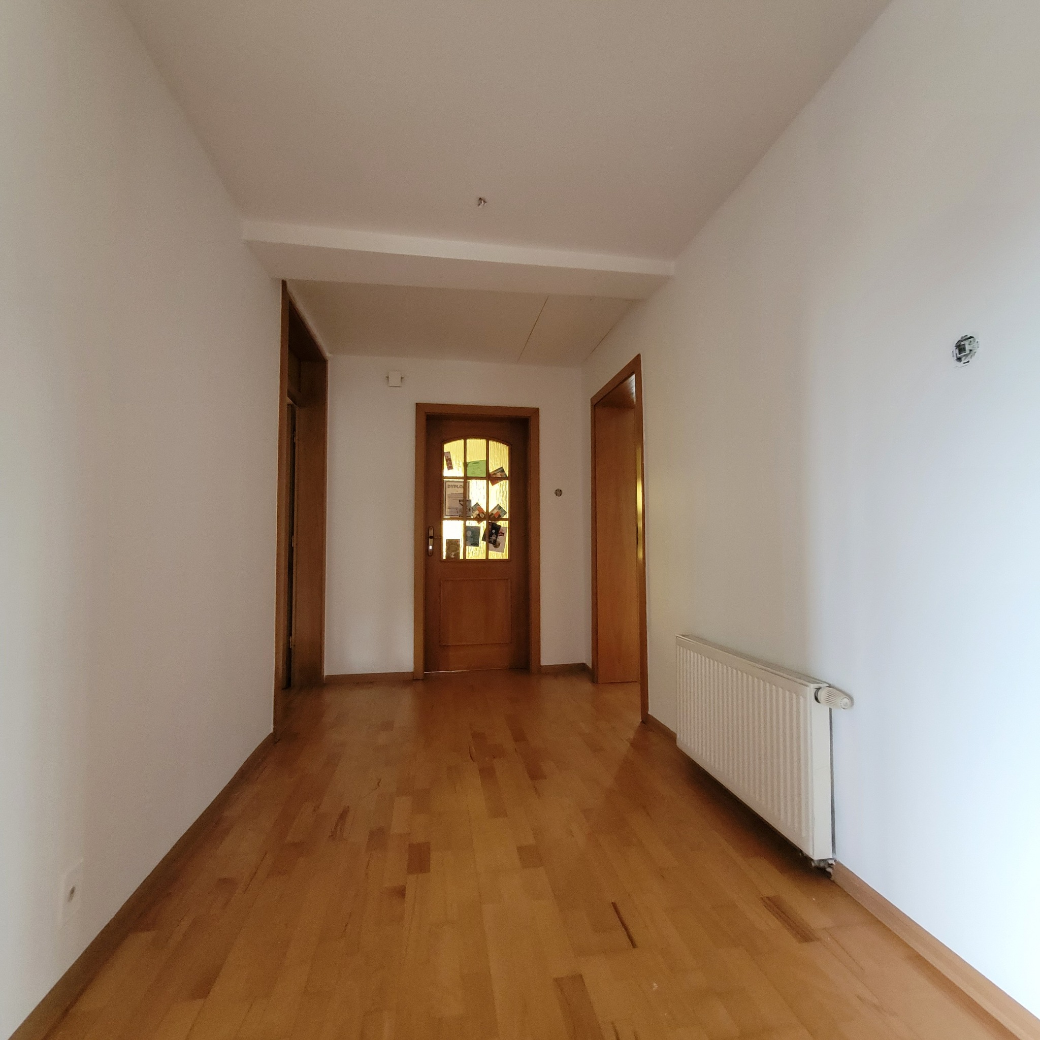 Puste, jasne wnętrze po odświeżeniu ścian, z drewnianą podłogą i grzejnikiem. Widoczna instalacja elektryczna bez osprzętu i drzwi z dyplomem.