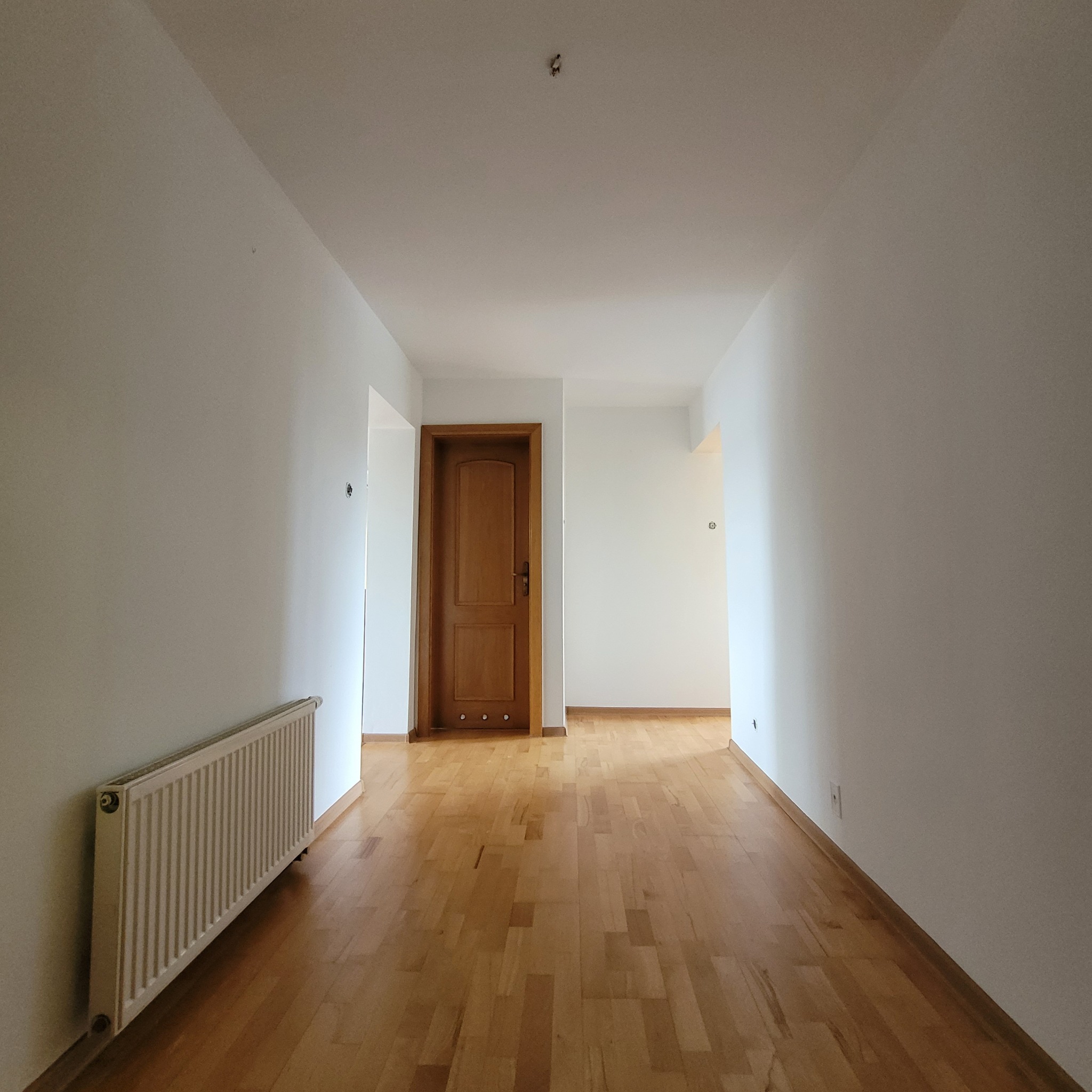 Białe ściany korytarza po odświeżeniu, z drewnianą podłogą i grzejnikiem. Widoczne gniazdka elektryczne i drewniane drzwi w głębi. Na suficie widoczny punkt świetlny.