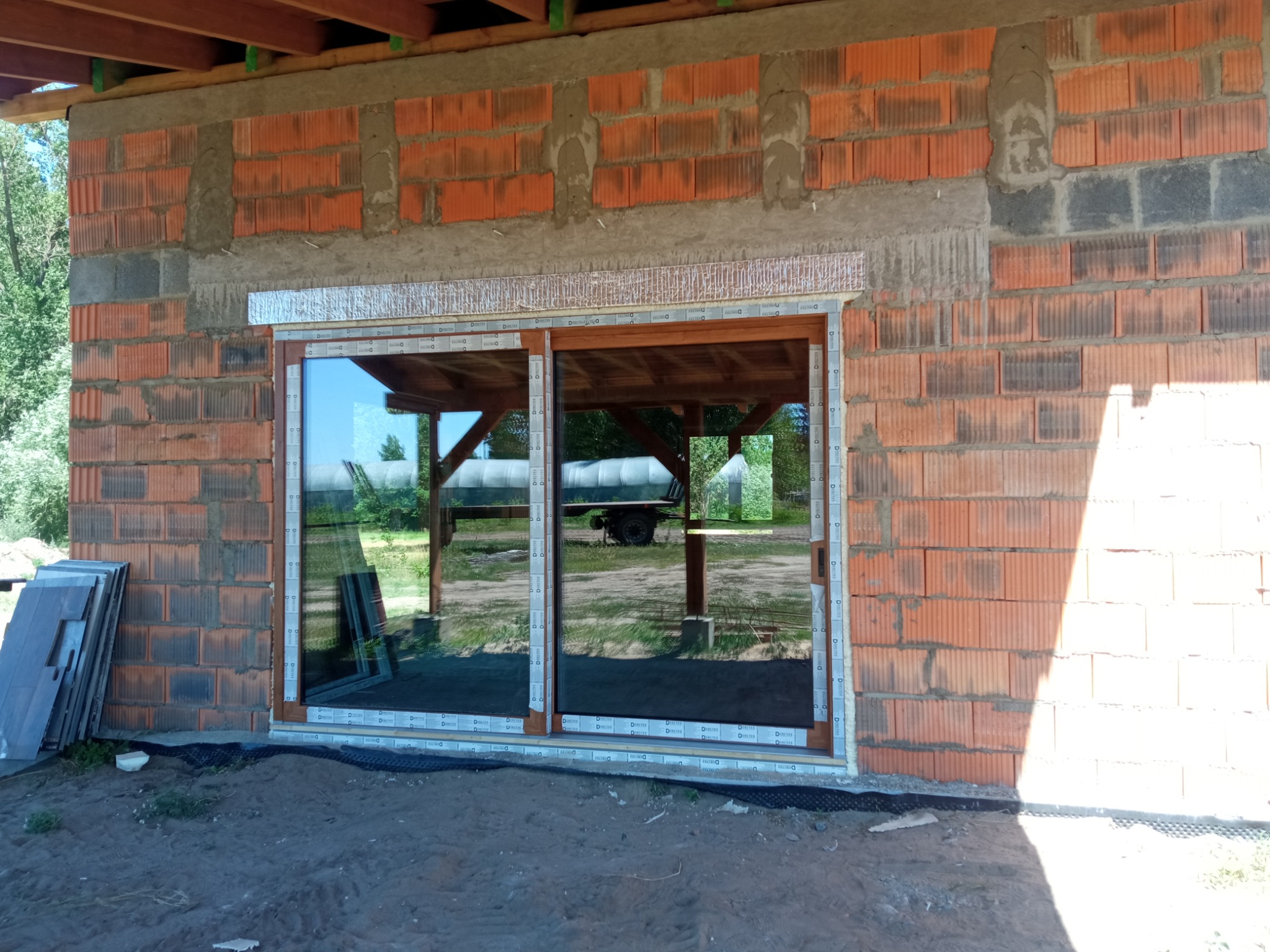 Montaż okna tarasowego z brązową ramą w surowym murze z cegły. Widoczne elementy konstrukcji dachu i przygotowane do montażu płyty. Przez szybę widać zieleń.