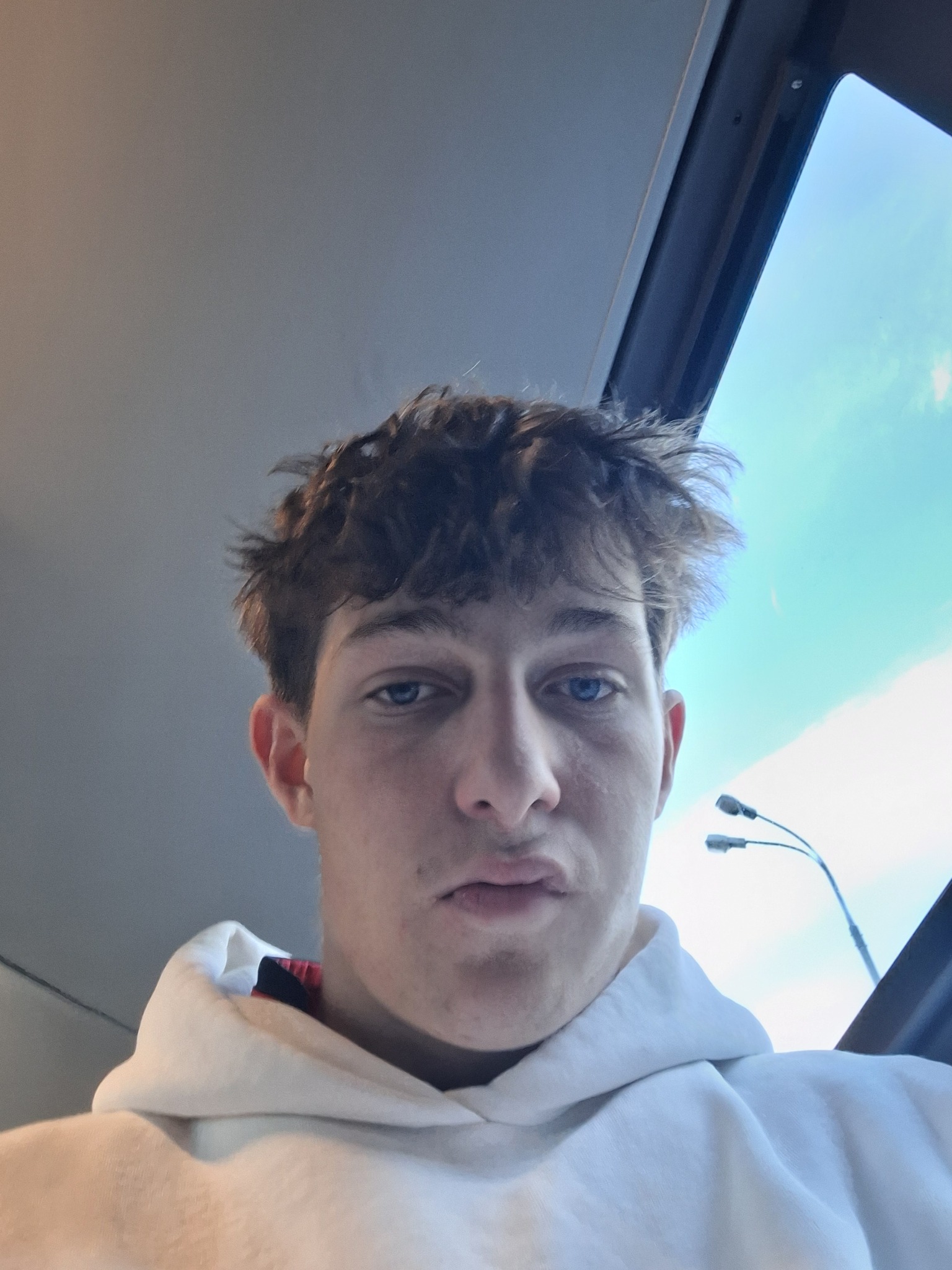 Portret młodego mężczyzny z niebieskimi oczami, patrzy w dół, ubrany w białą bluzę z kapturem. W tle widoczne niebo i fragment ulicznej lampy.