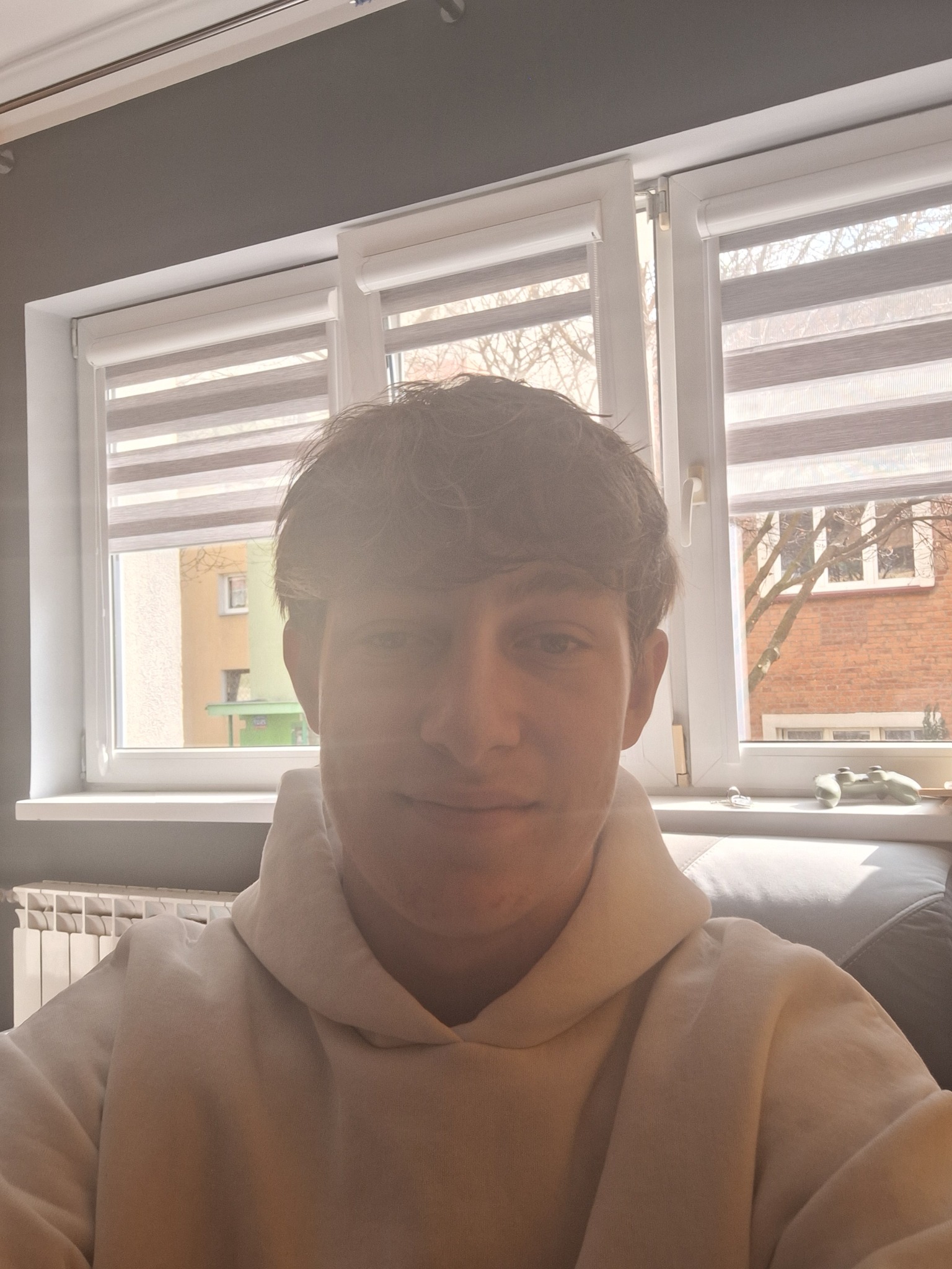 Portret młodego mężczyzny w jasnej bluzie z kapturem na tle okna z roletami. Na parapecie widoczny kontroler do gier. Ujęcie z bliska, naturalne światło.