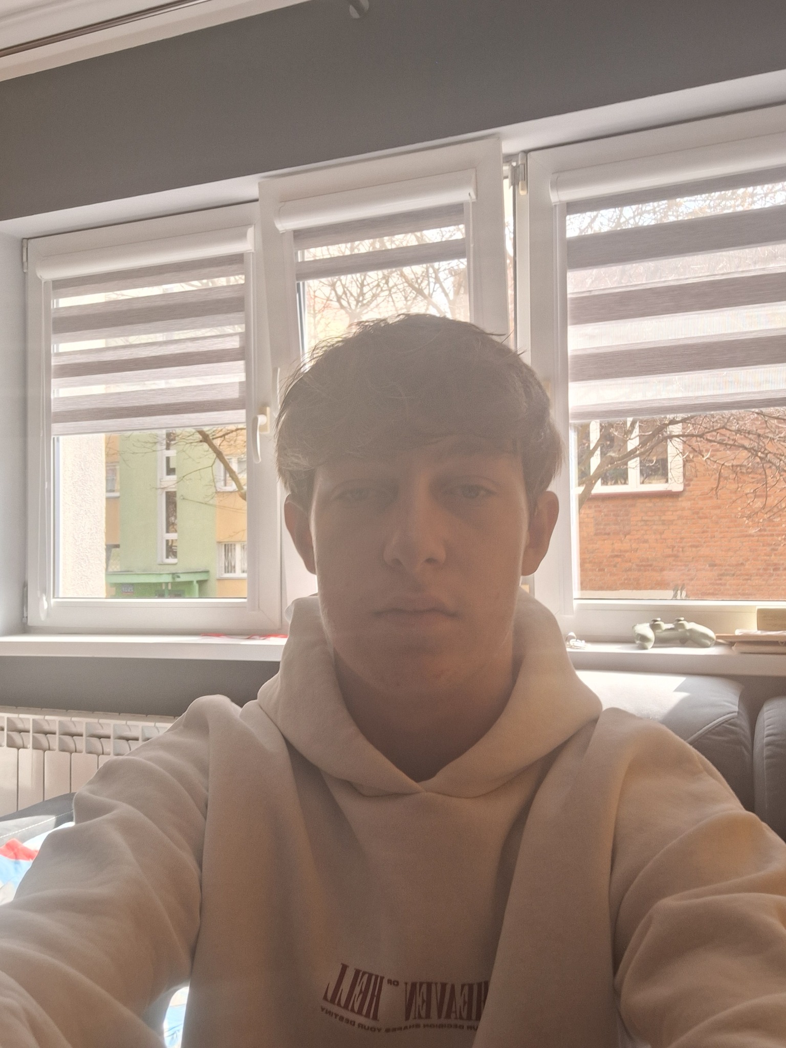 Młody mężczyzna w białej bluzie z kapturem, selfie w jasnym pokoju z żaluzjami i widokiem na budynki. Na parapecie leży biały kontroler do gier.