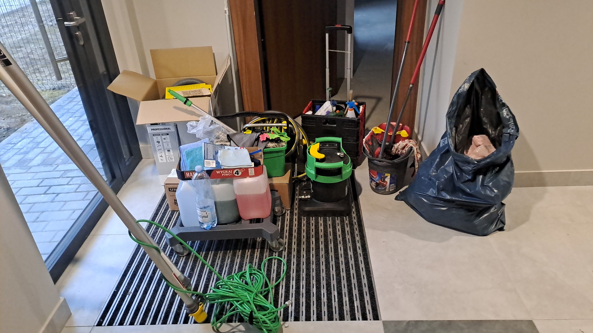 Sprzęt i akcesoria do sprzątania: wózek z płynami, odkurzacz, mop, wiadro i worek na śmieci, gotowe do użycia w pomieszczeniu z kratką podłogową.