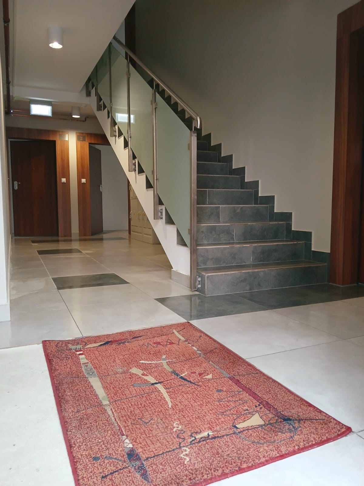 Wnętrze budynku z klatką schodową o szklanych balustradach i ciemnych stopniach. Na podłodze leży wzorzysty, czerwony dywan. Widoczne drzwi i fragmenty podłogi.