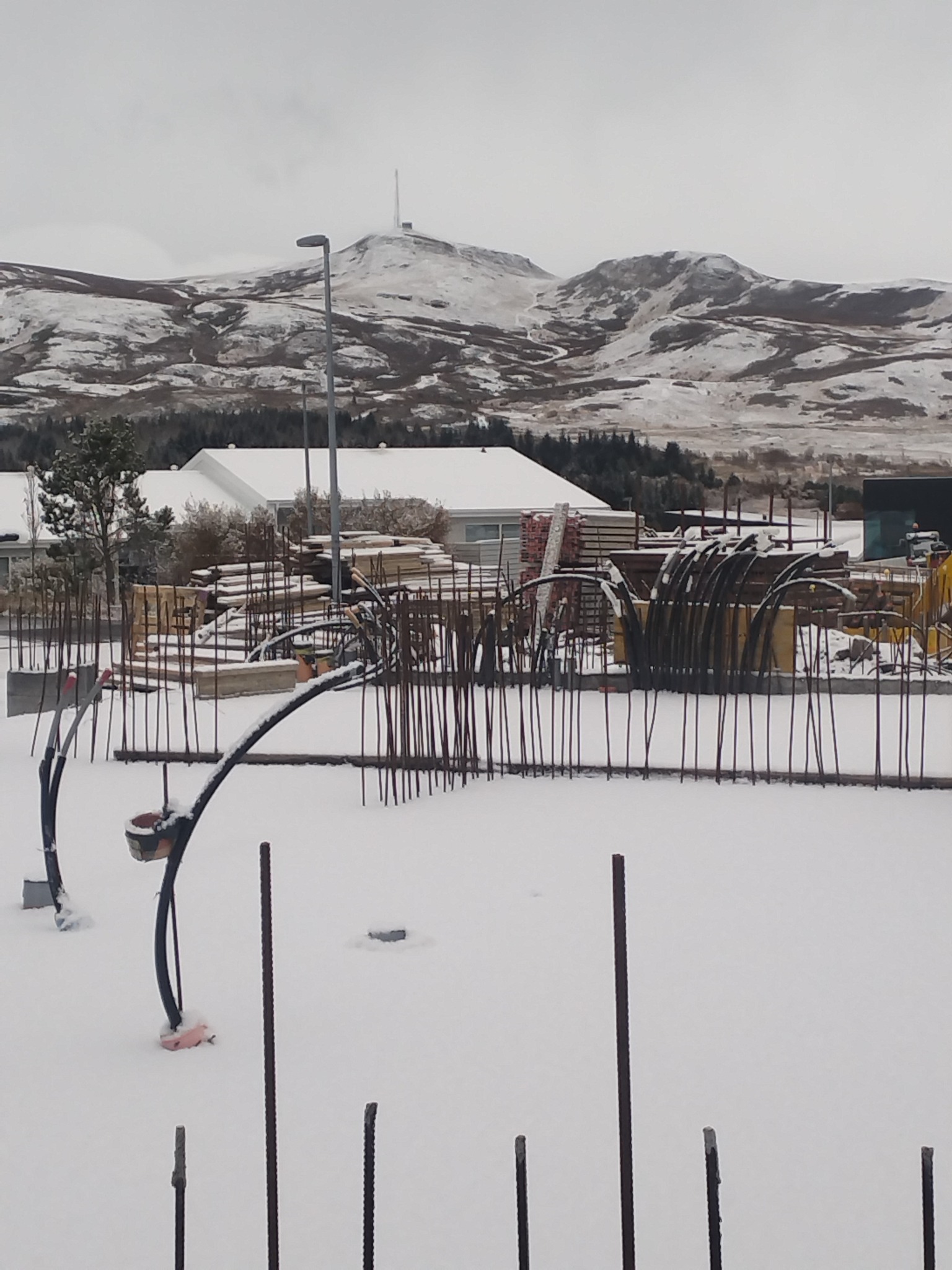 Zimowy krajobraz z placem budowy: wystające pręty zbrojeniowe, drewniane elementy konstrukcyjne pod śniegiem, w tle góry i budynki.