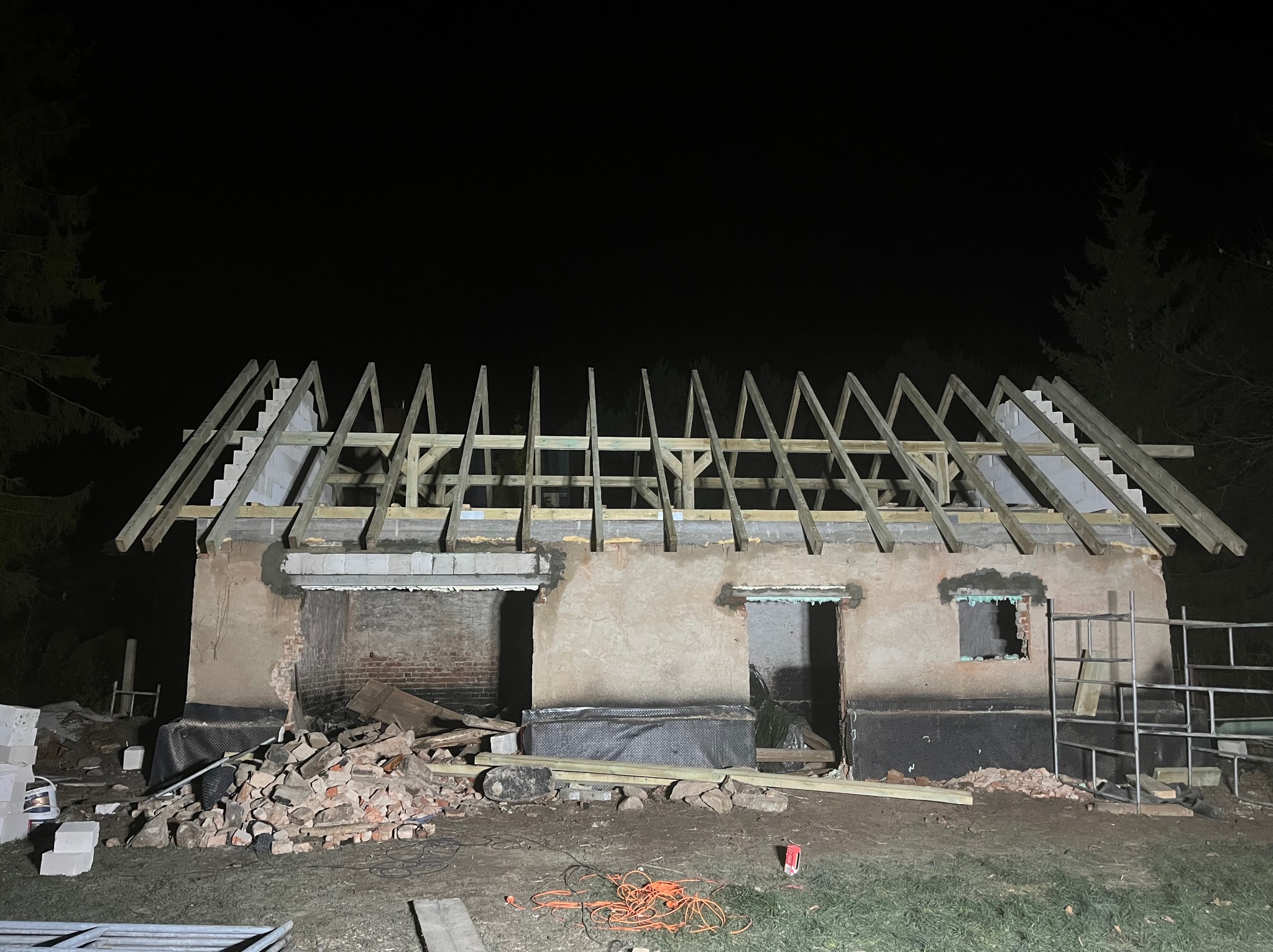 Nocne ujęcie wznoszonej konstrukcji dachu z drewnianych belek na murowanym budynku w stanie surowym otwartym, z gruzem i narzędziami na pierwszym planie.