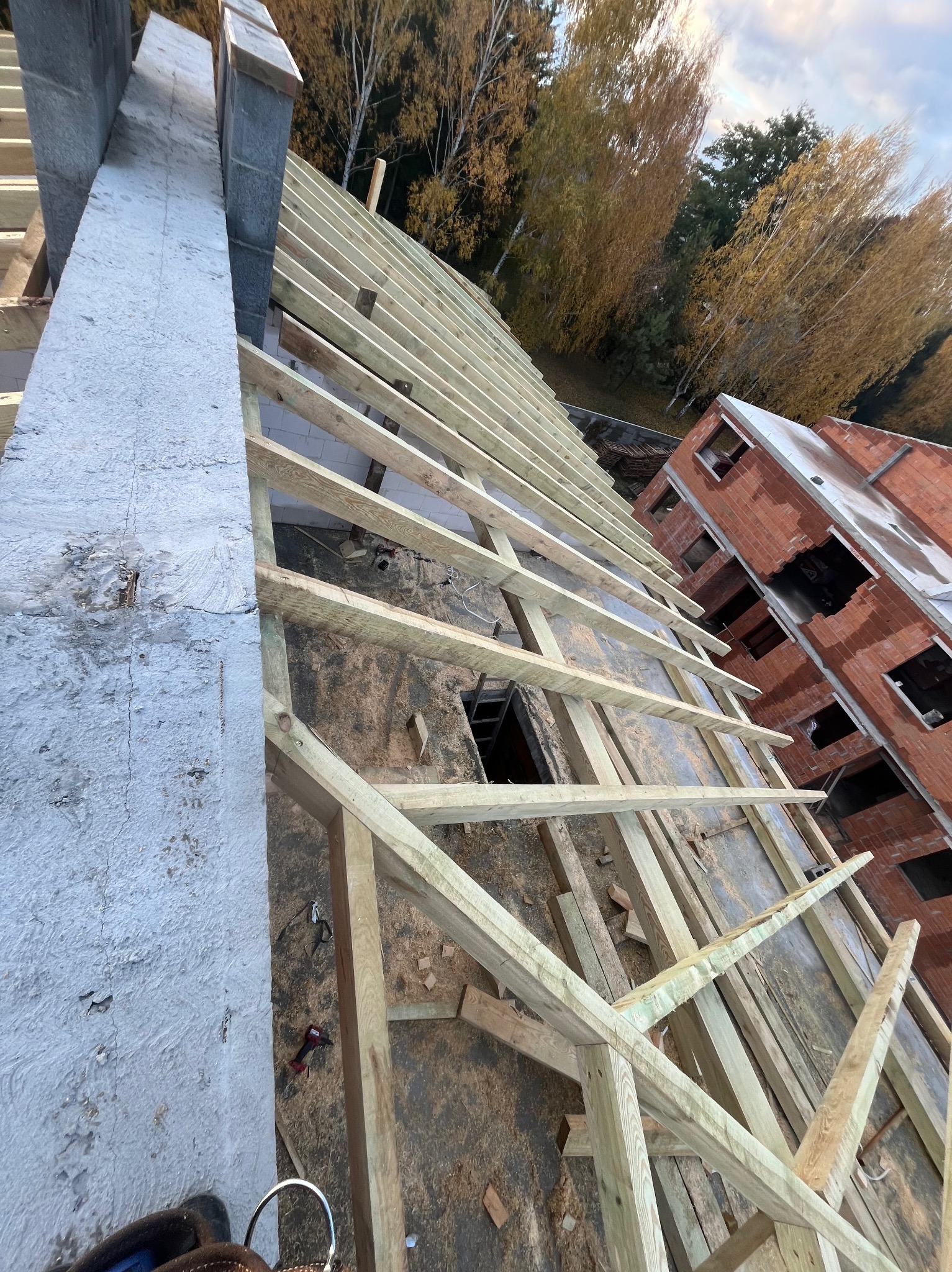 Konstrukcja drewniana dachu w trakcie budowy, widok z góry, z fragmentem murowanej ściany i jesiennym lasem w tle. Widoczne narzędzia pracy.