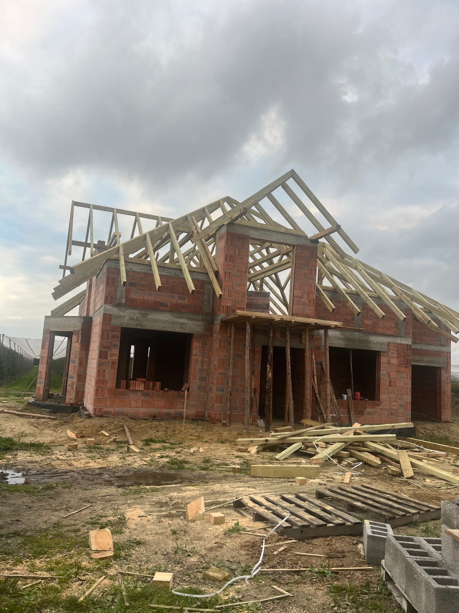 Budowa domu jednorodzinnego w trakcie wznoszenia. Widoczna konstrukcja dachu z drewnianych belek na tle pochmurnego nieba. Na pierwszym planie porozrzucane deski i bloczki.