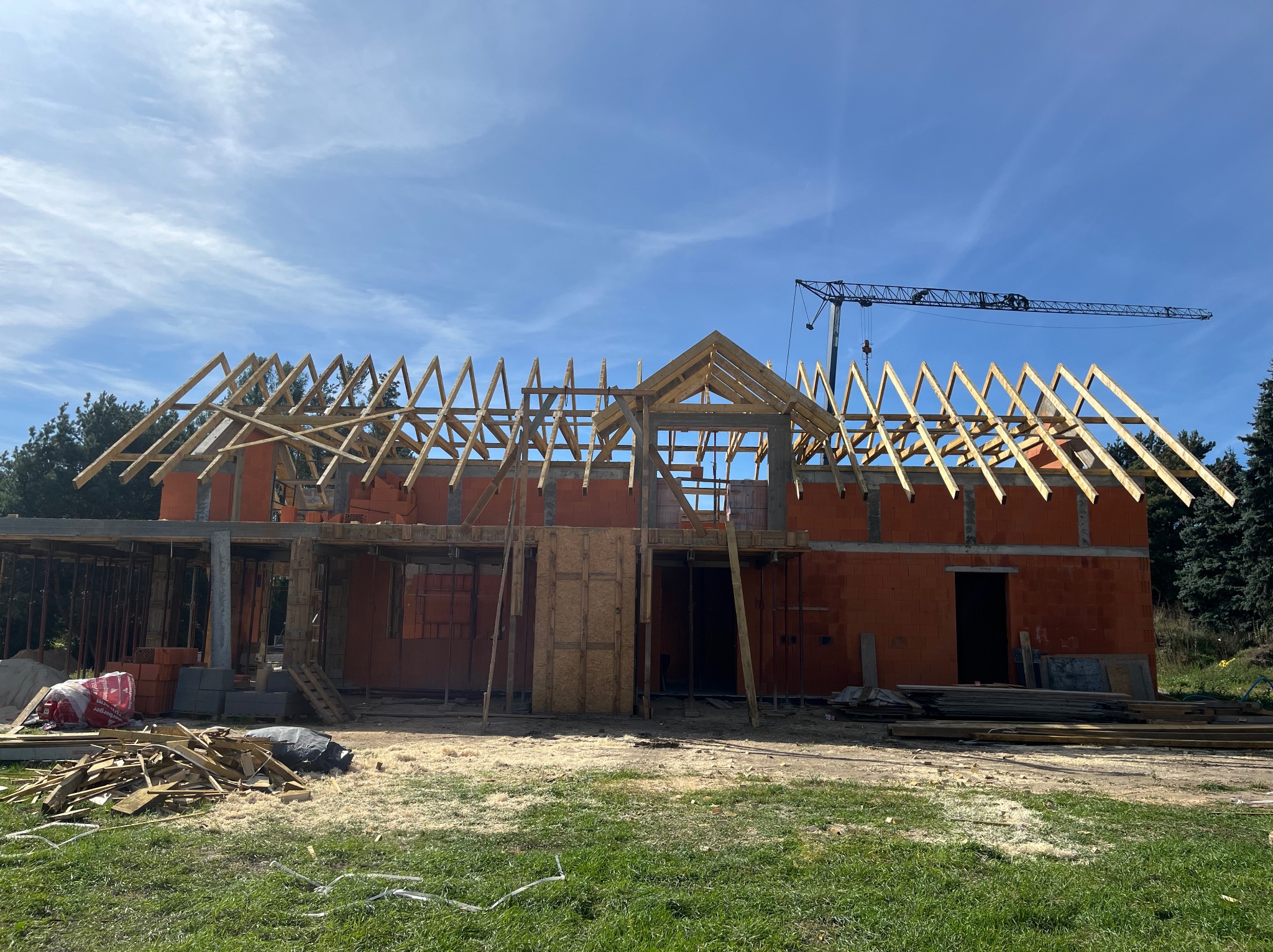 Budowa domu z cegły: wzniesiona konstrukcja ścian i drewniana konstrukcja dachu w trakcie montażu. W tle widoczny żuraw budowlany i błękitne niebo.