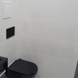 Nowoczesna toaleta z czarną armaturą i białymi płytkami. Minimalistyczny design, czarny sedes i umywalka. W tle widoczne białe płytki i czarny przycisk spłuczki.