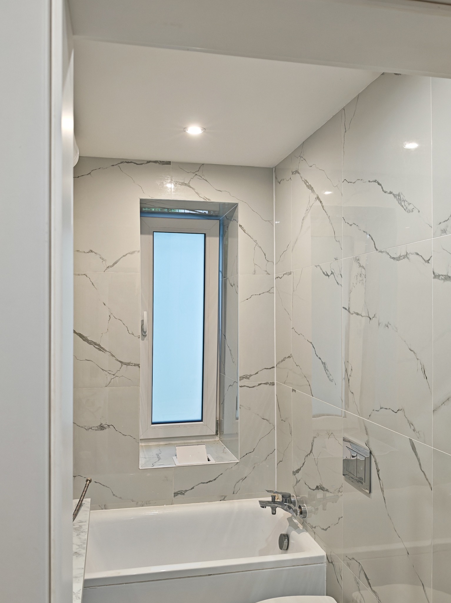 Biała łazienka z wanną i oknem, wykończona płytkami imitującymi marmur z szarymi żyłami. Armatura chromowana, oświetlenie punktowe.