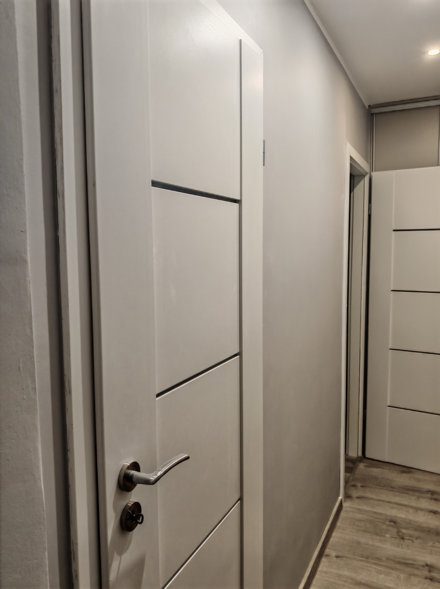 Nowoczesne, białe drzwi wewnętrzne z poziomymi frezami i srebrną klamką, widoczne w korytarzu z drewnopodobną podłogą i szarą ścianą, w tle kolejne drzwi.