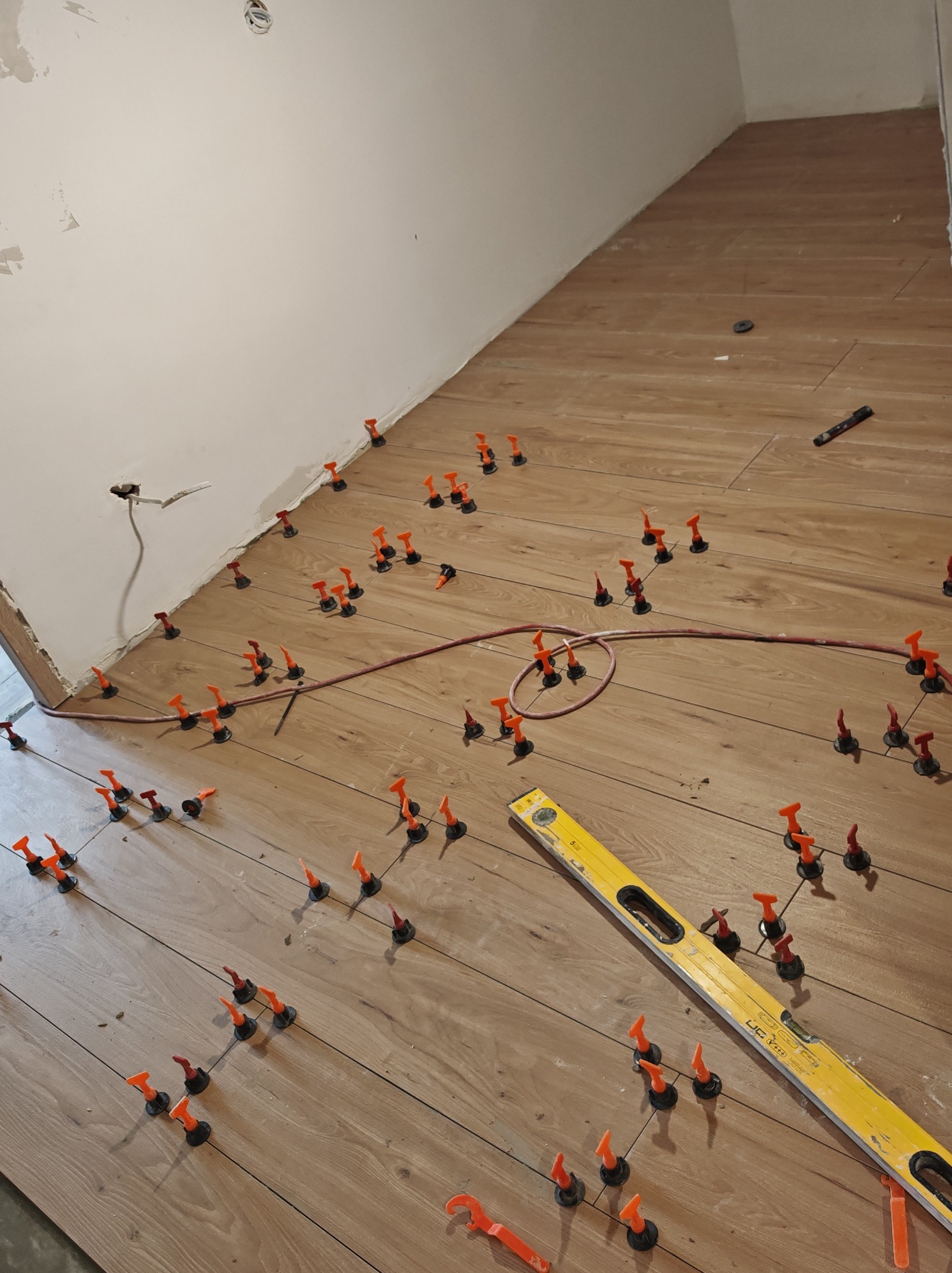 Układanie paneli podłogowych z użyciem klipsów poziomujących; widoczna poziomica i ściana w trakcie prac wykończeniowych. Jasne panele imitujące drewno.