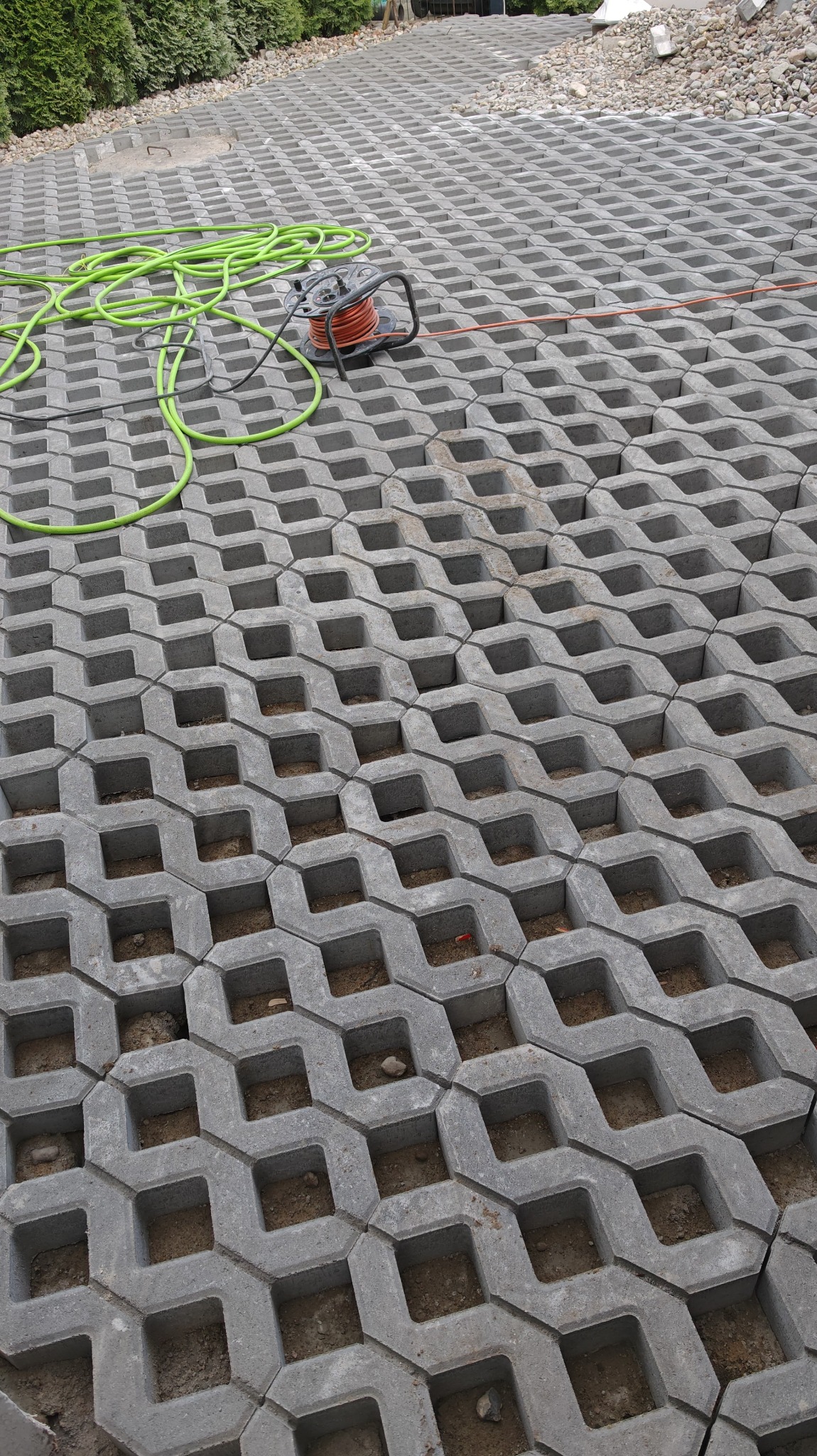 Ułożona szara ażurowa kostka brukowa na podjeździe, z widocznym zielonym wężem ogrodowym i zwiniętym przedłużaczem elektrycznym. Widok z góry.