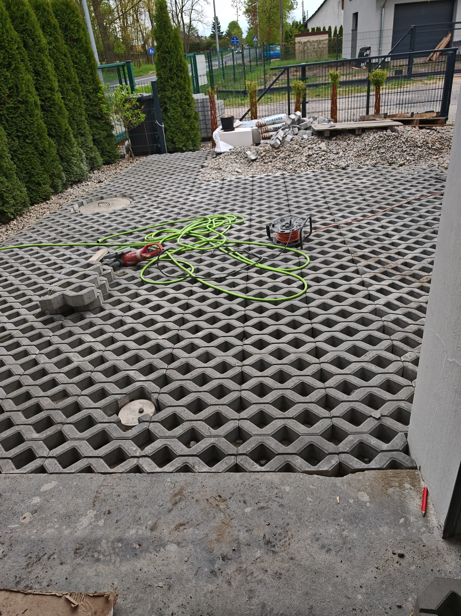 Układanie ażurowej kostki brukowej na podjeździe. Widoczny fragment ułożonej nawierzchni, narzędzia i materiały budowlane. Tło stanowią tuje i brama wjazdowa.