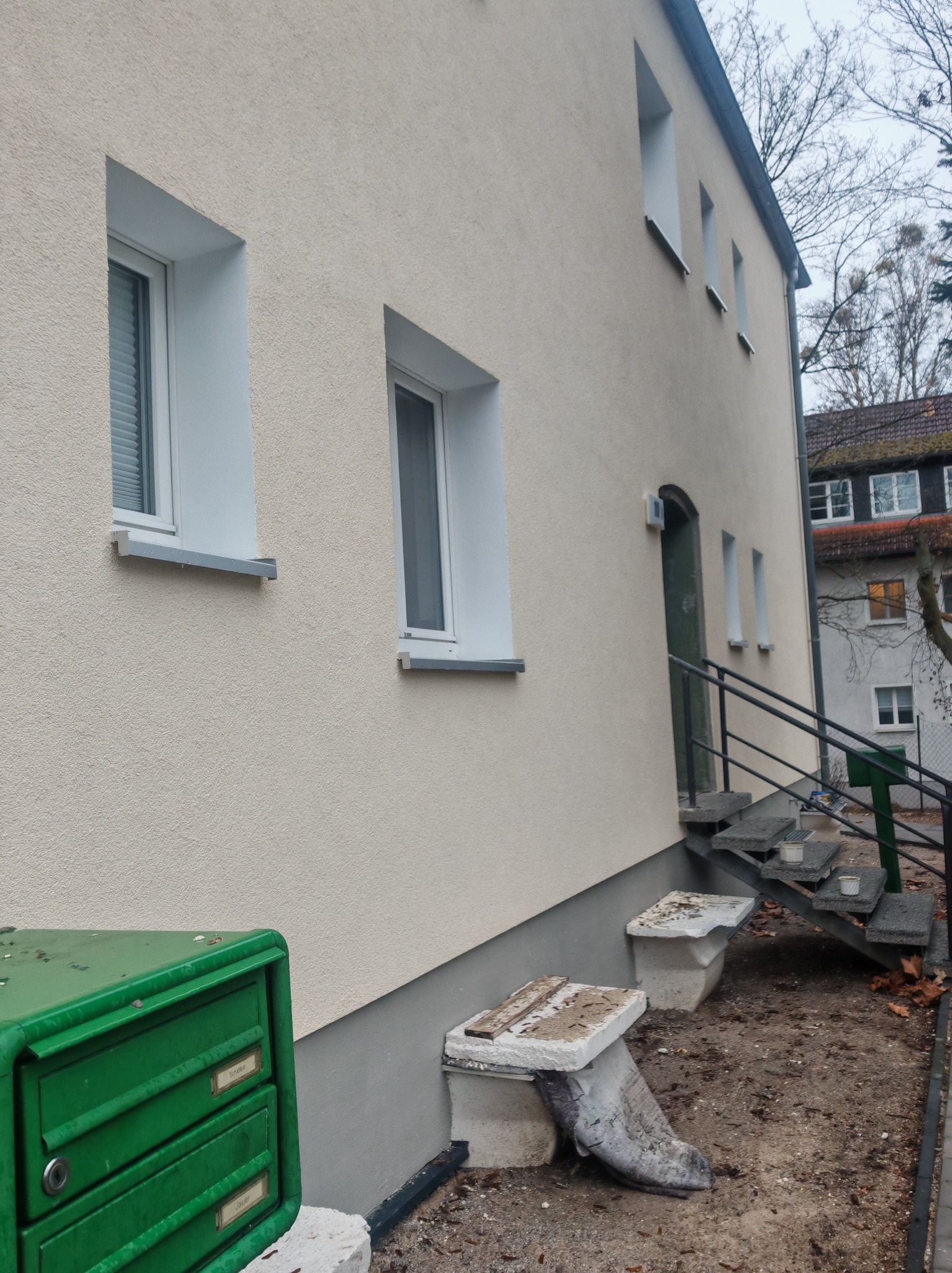 Odnowiona elewacja budynku wielorodzinnego z nowymi oknami i zieloną skrzynką pocztową. Widoczne schody wejściowe z metalową barierką.