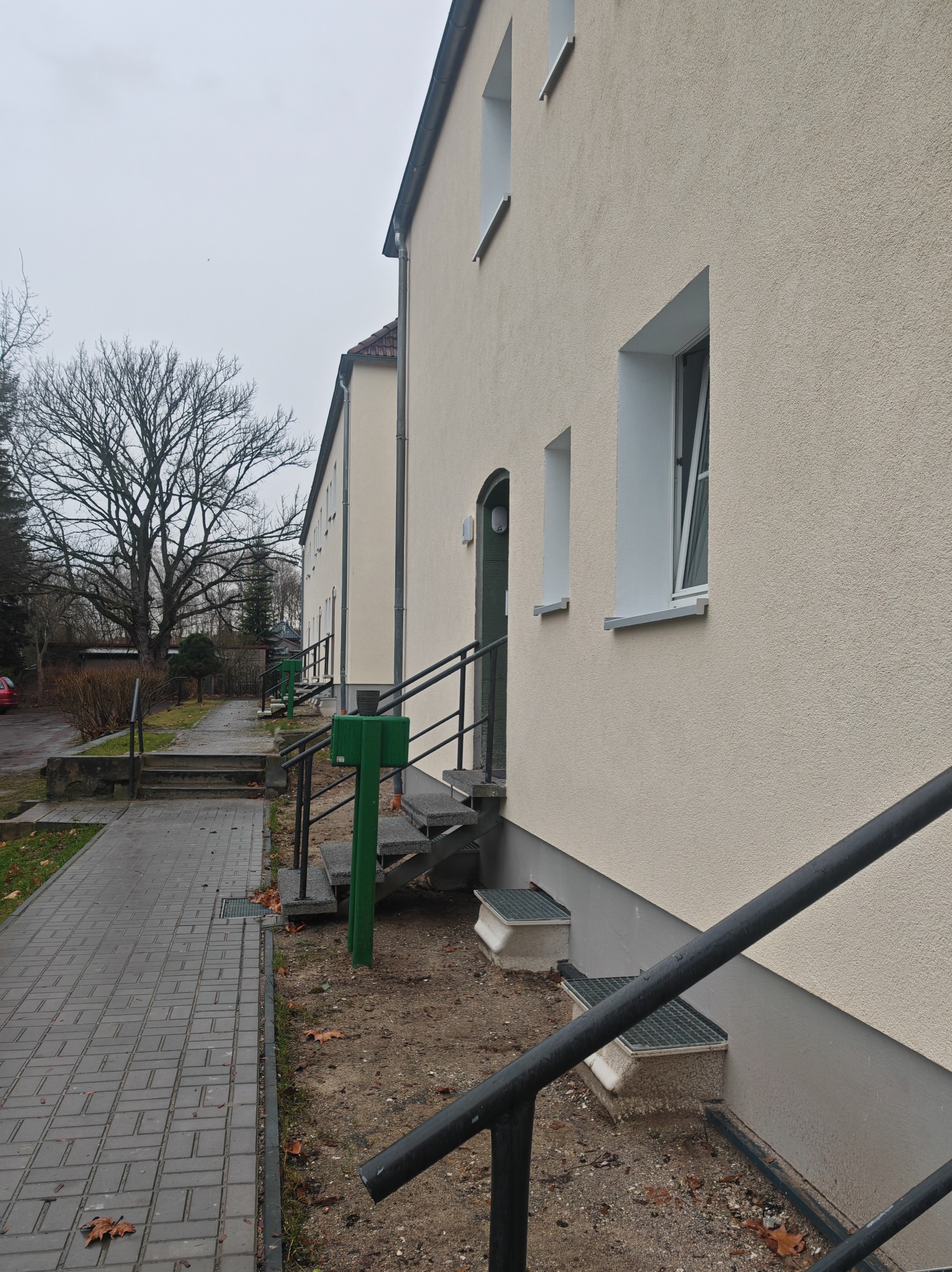 Elewacja budynku po tynkowaniu i malowaniu, widoczne okna z białymi ramami, wejście z zielonymi drzwiami i metalowymi schodami. Na pierwszym planie fragment chodnika.