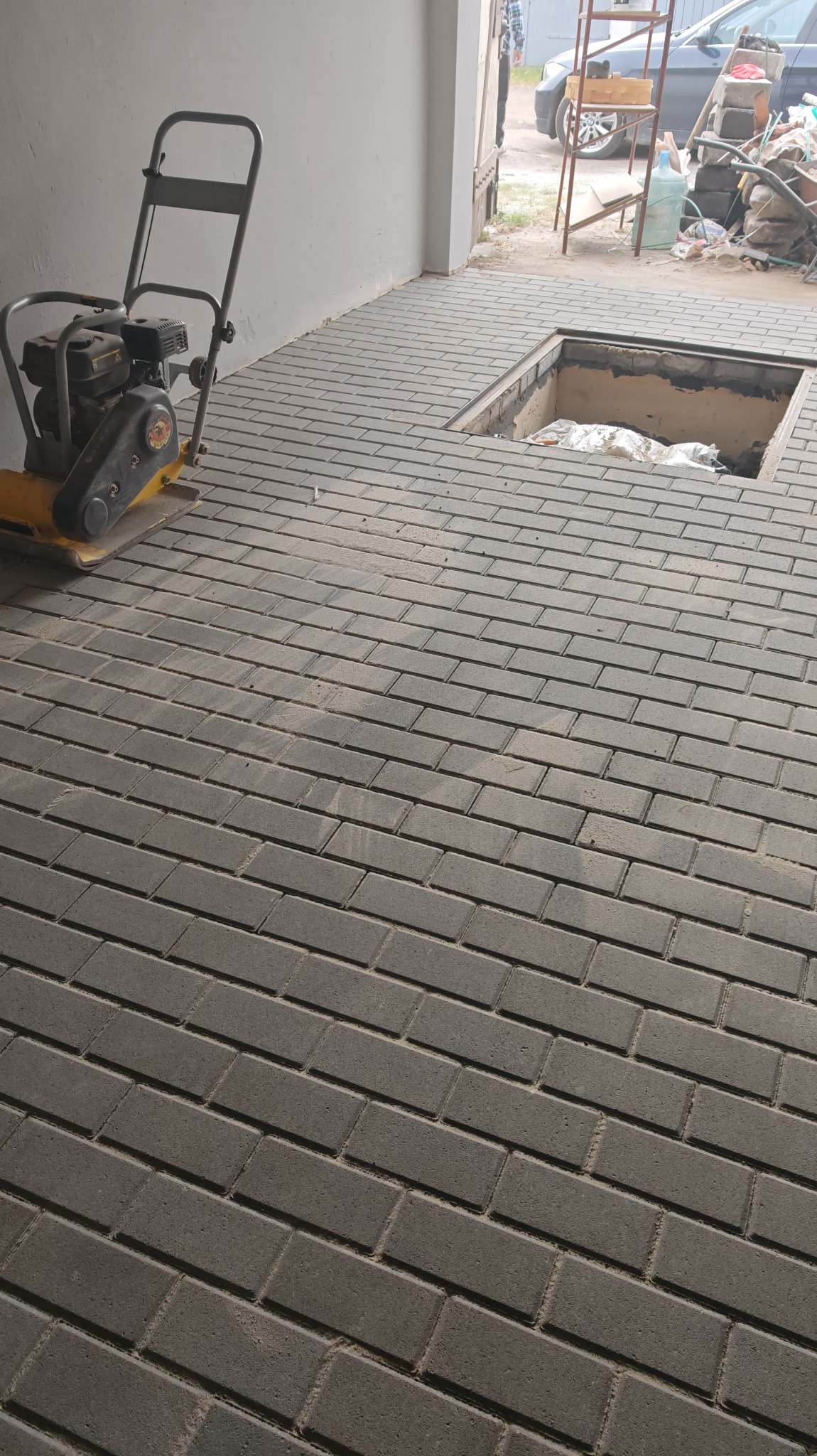 Szara kostka brukowa ułożona na podjeździe, z widocznym otworem inspekcyjnym i zagęszczarką w tle. Detal wykończenia, praca brukarska w trakcie.