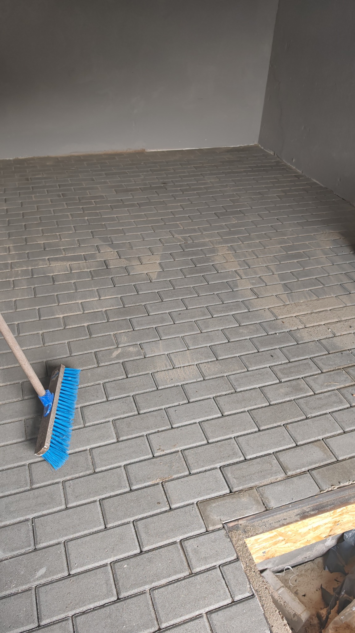 Ułożona szara kostka brukowa w Kostrzynie nad Odrą. Część powierzchni posprzątana szczotką z niebieskim włosiem. Widoczny fragment ściany i krawędź wykończenia.